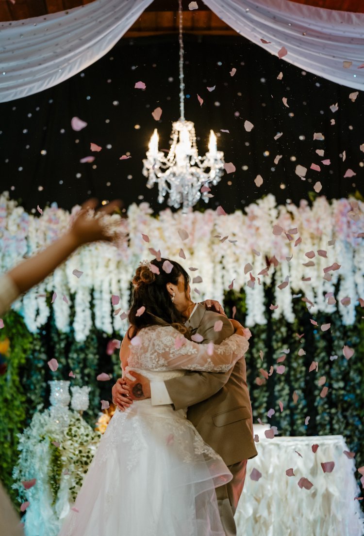 A couple embracing at a wedding with pink petals falling, under a chandelier and floral decorations.
