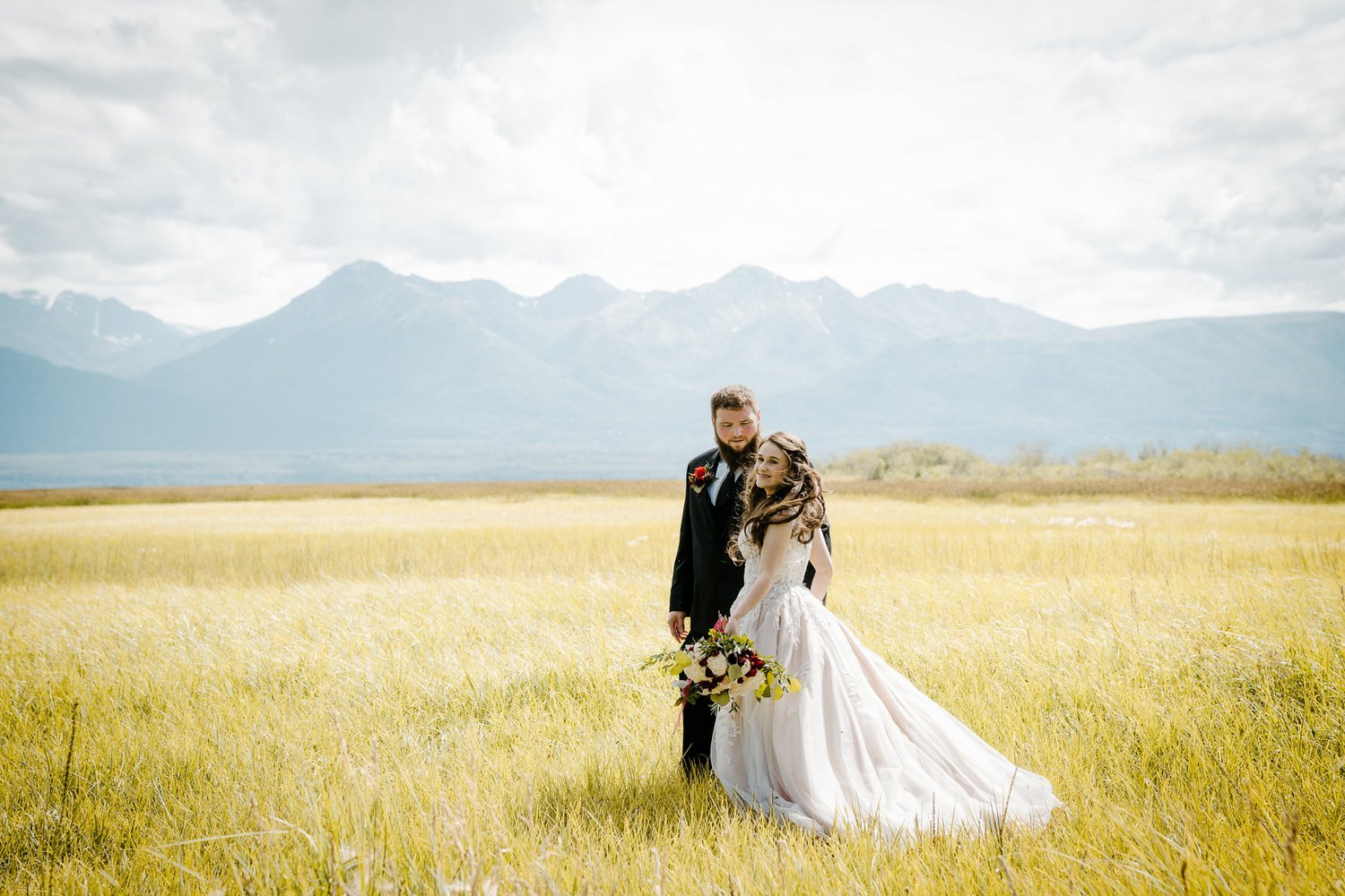 Bride and groom standing in a grassy field with mountains in the background.