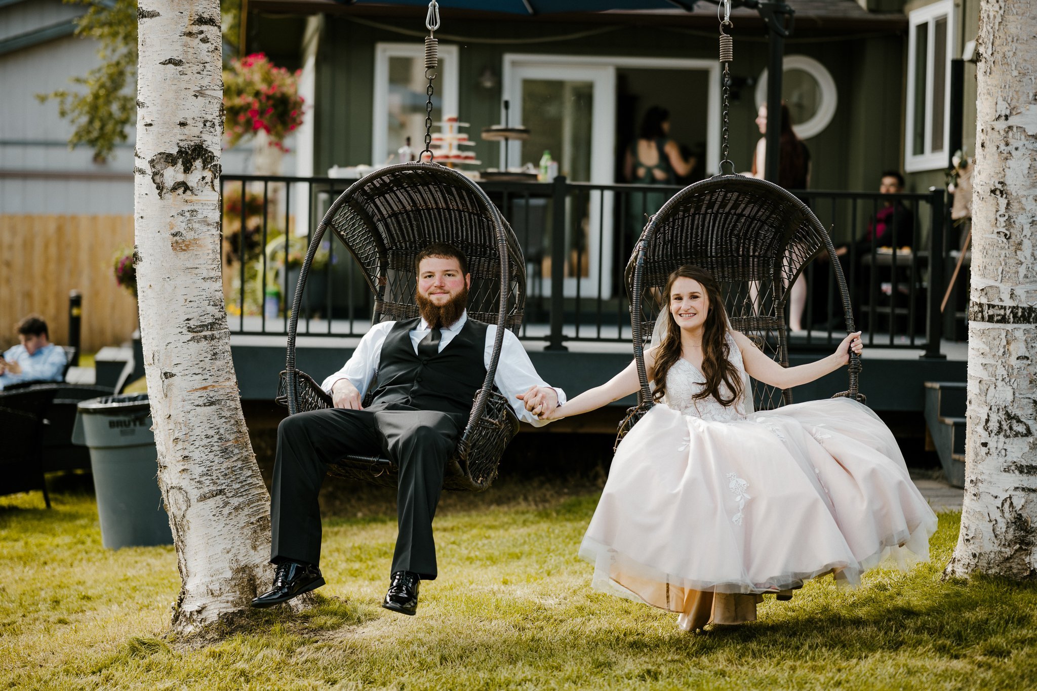 A couple in formal attire sitting on hanging chairs outdoors, holding hands. The man is wearing a black suit, and the woman is wearing a white wedding dress. They are in a grassy area with a porch and people in the background.