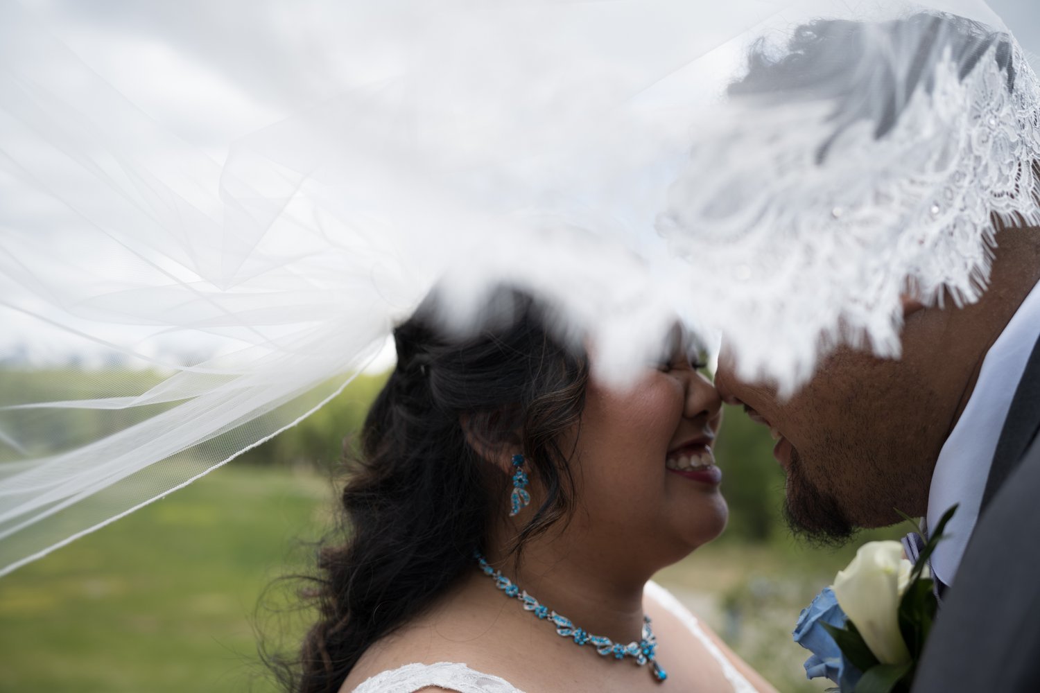 Bride and groom smiling under a billowing wedding veil with close proximity, bride wearing blue jewelry, groom in a suit holding flowers.
