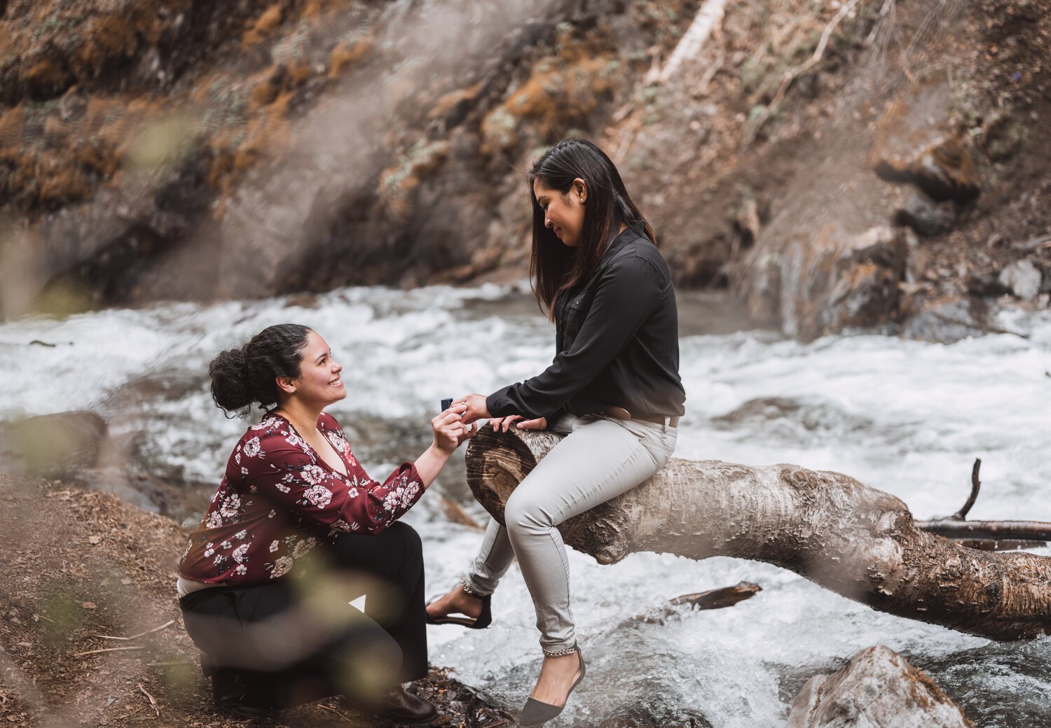 A woman proposing to another woman by a river in a forested area.