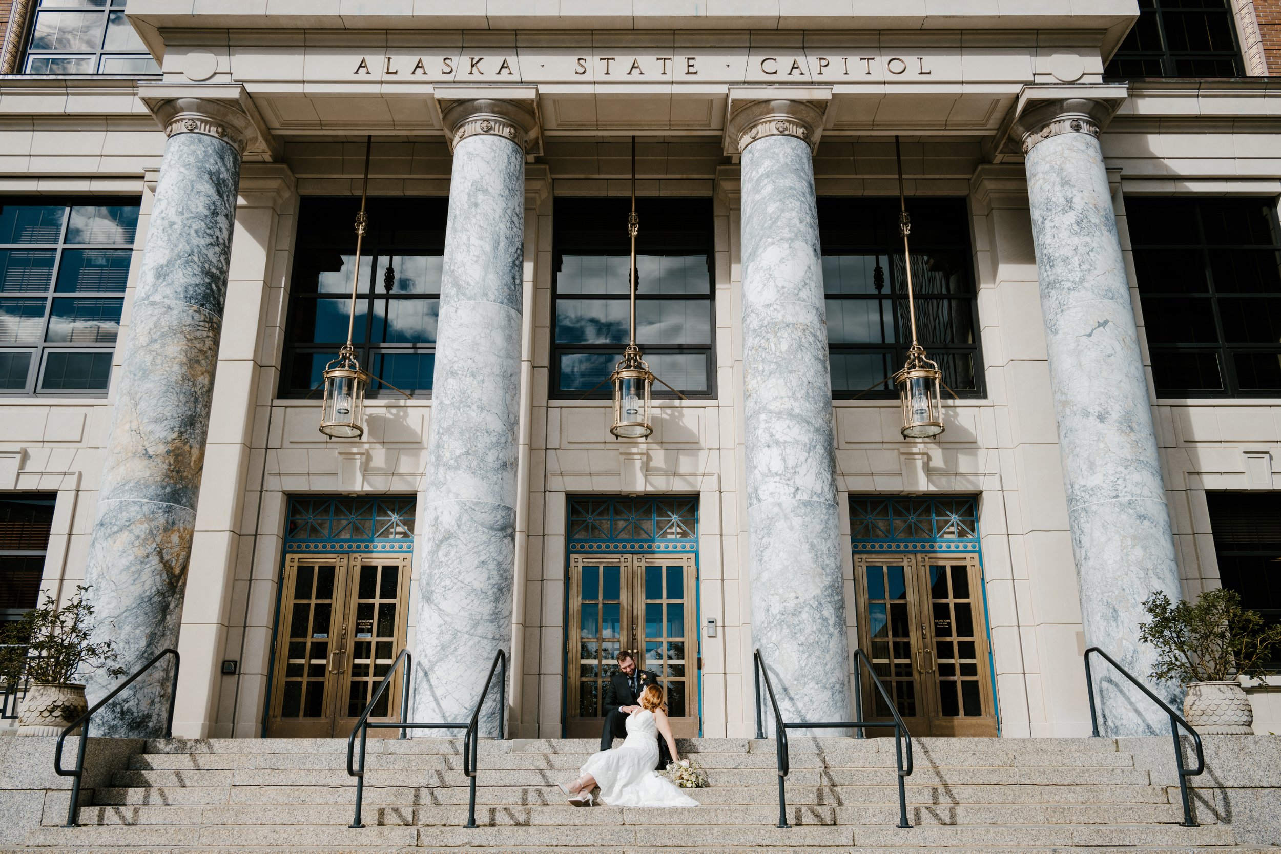 Bride and groom posing in front of Alaska State Capitol building with large columns and entrance steps.