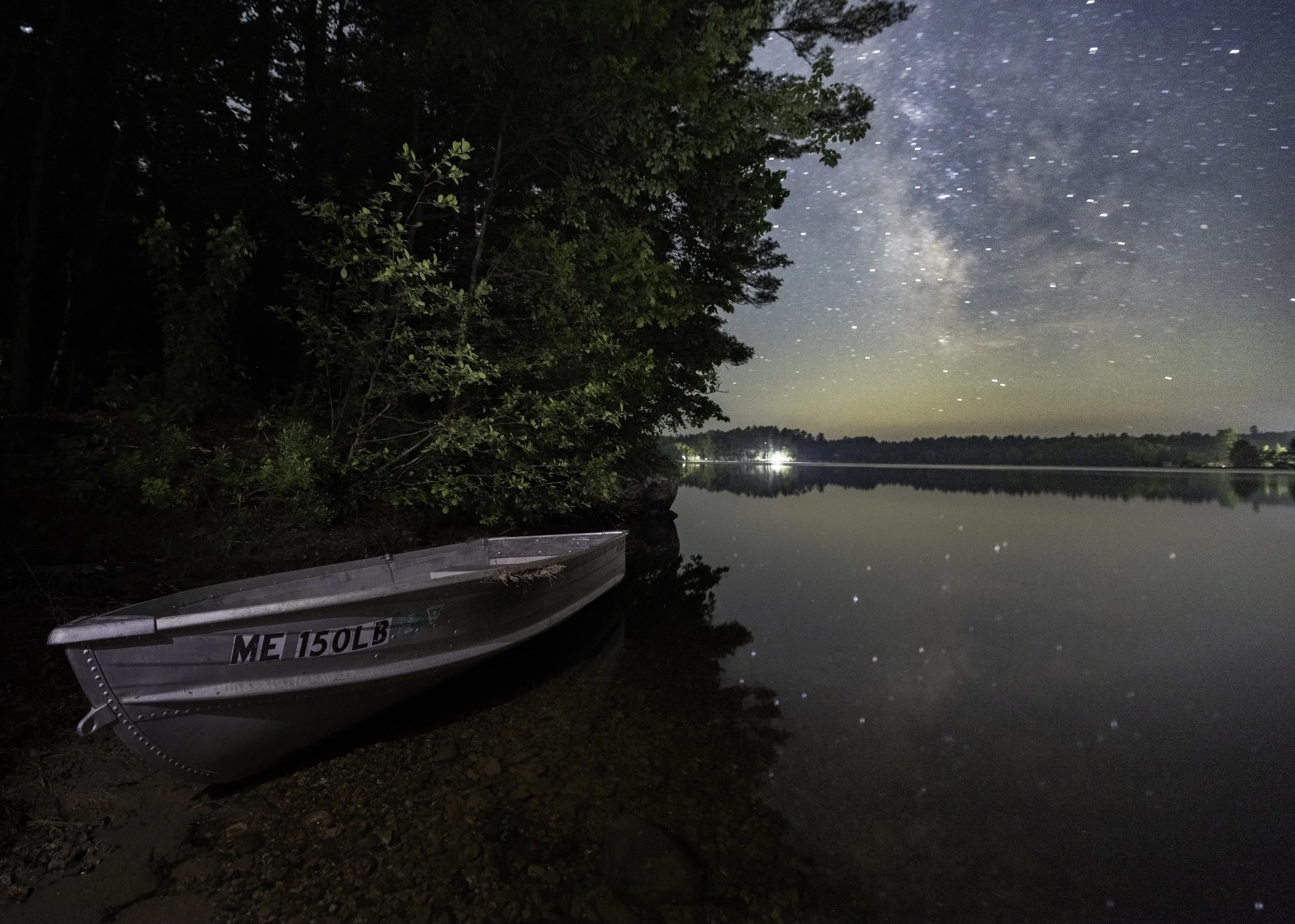 A small rowboat resting on a rocky shoreline at night, next to dense trees, under a starry sky and with a calm reflective lake surface.