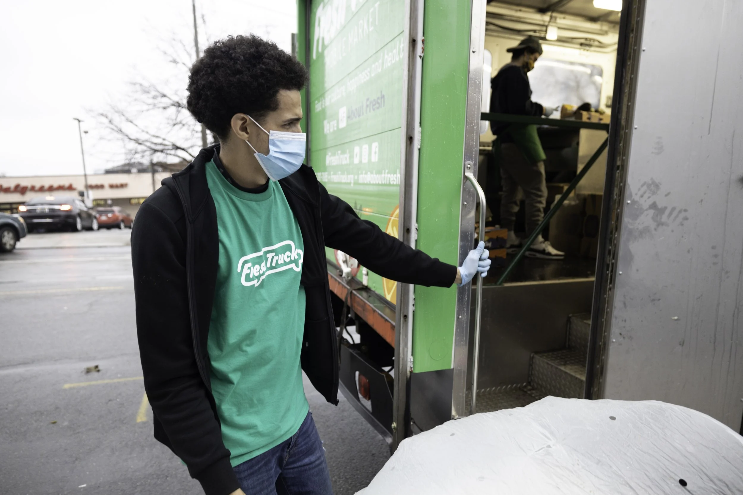 A young man wearing a face mask and a teal shirt with 'FastTrak' logo unloading from a green food truck