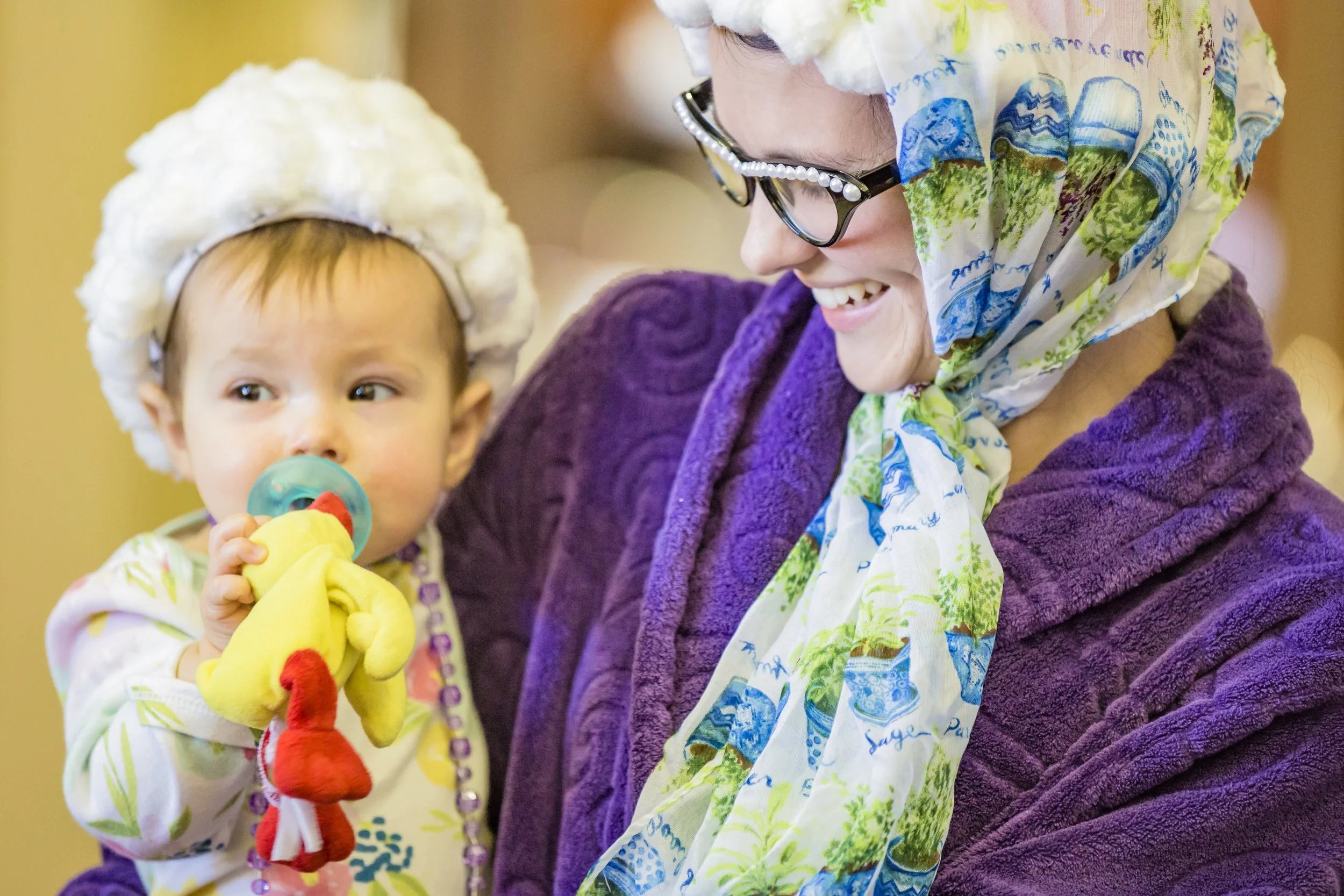 A woman smiling and wearing glasses, a purple coat, and a colorful scarf, holding a young child with a pacifier, a plush chicken toy, and a fluffy white hat.