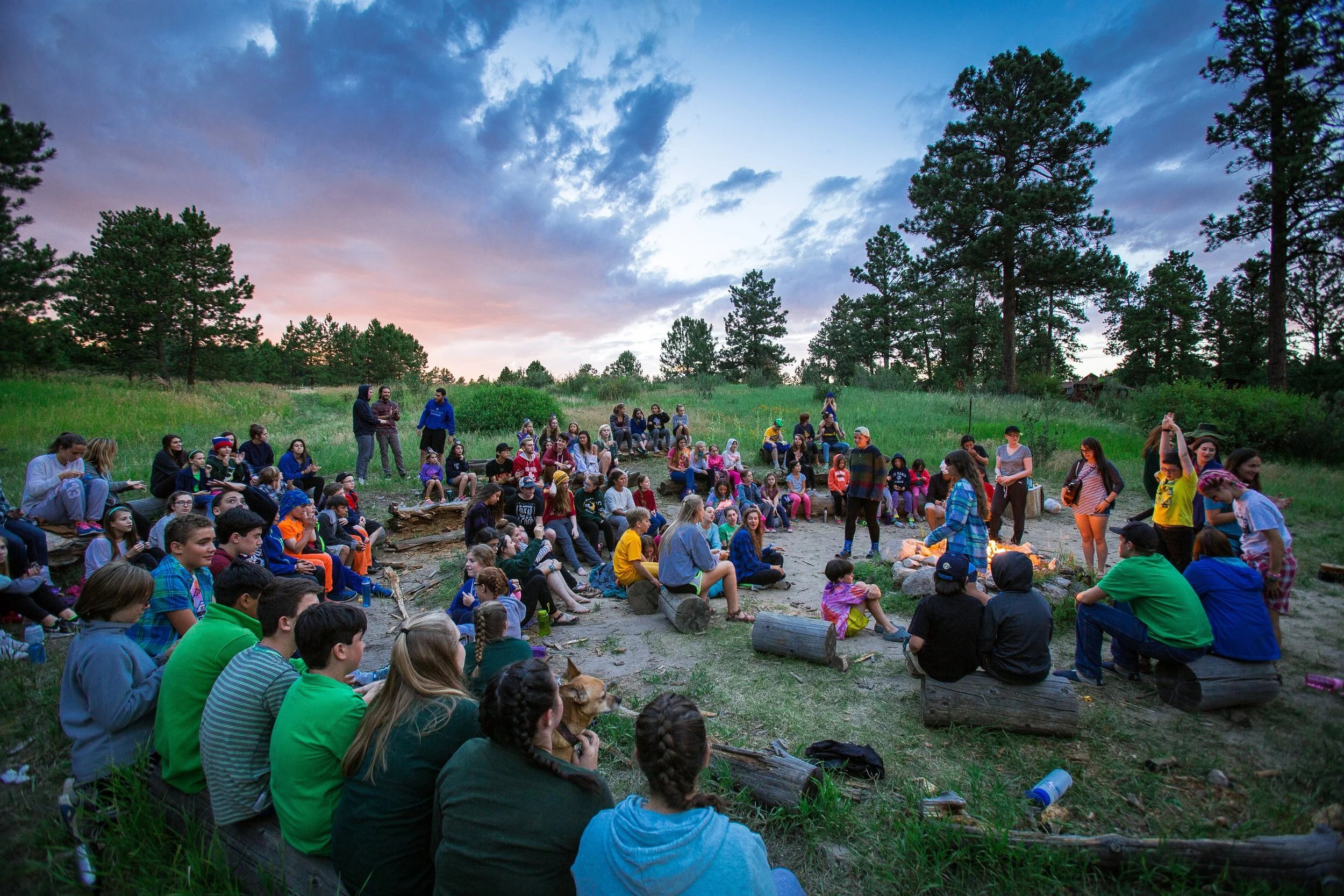 A large group of children and adults gathered outdoors around a campfire during sunset in a forested area.