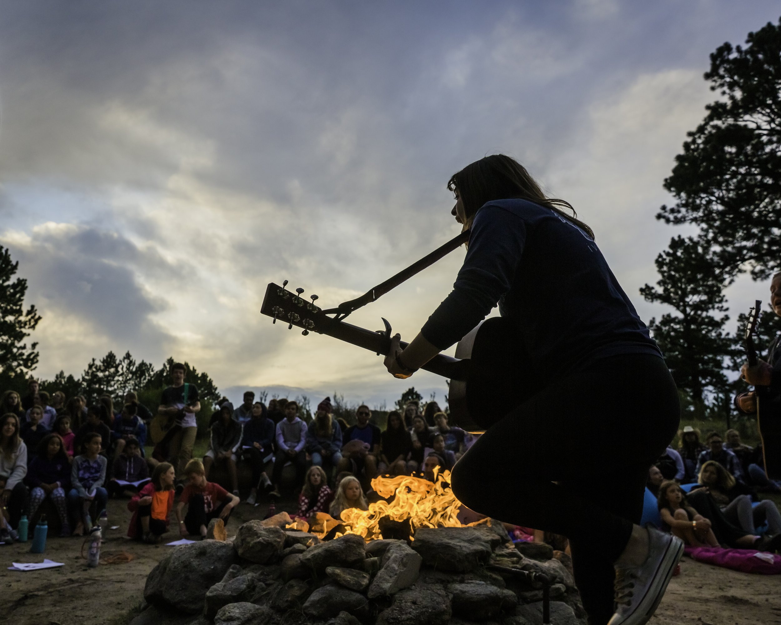 A woman playing guitar near a campfire outdoors during sunset or twilight with a crowd of people sitting on rocks and ground and trees in the background.