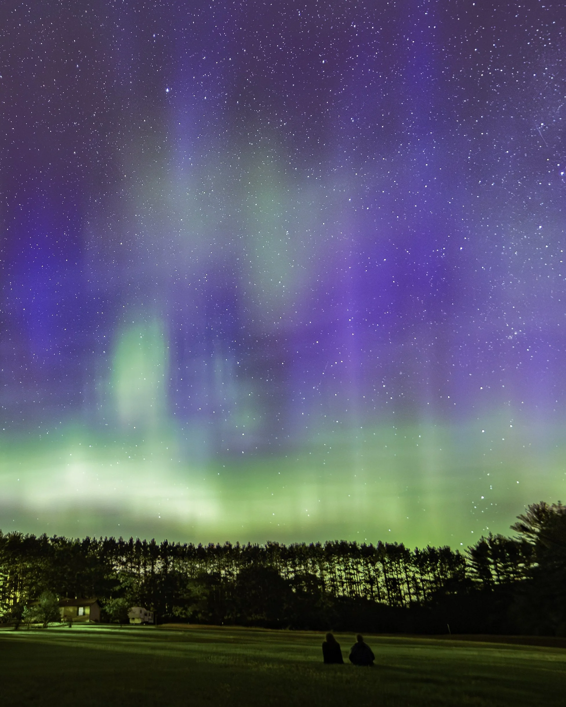 Two people sitting on a grassy field, watching the northern lights in the night sky filled with stars and colorful auroras.