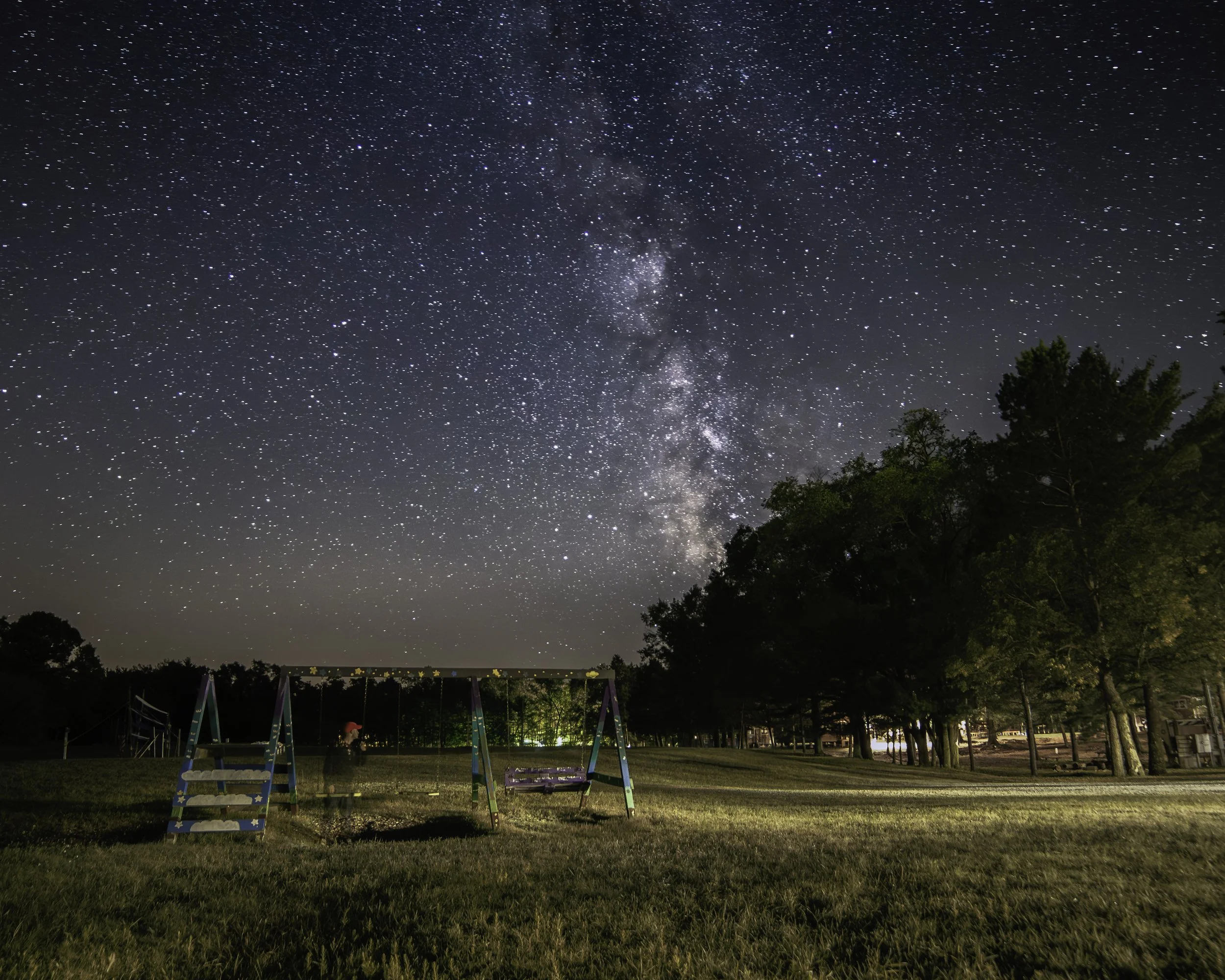 Nighttime scene at a park with swings, illuminated trees, and a star-filled sky with the Milky Way.