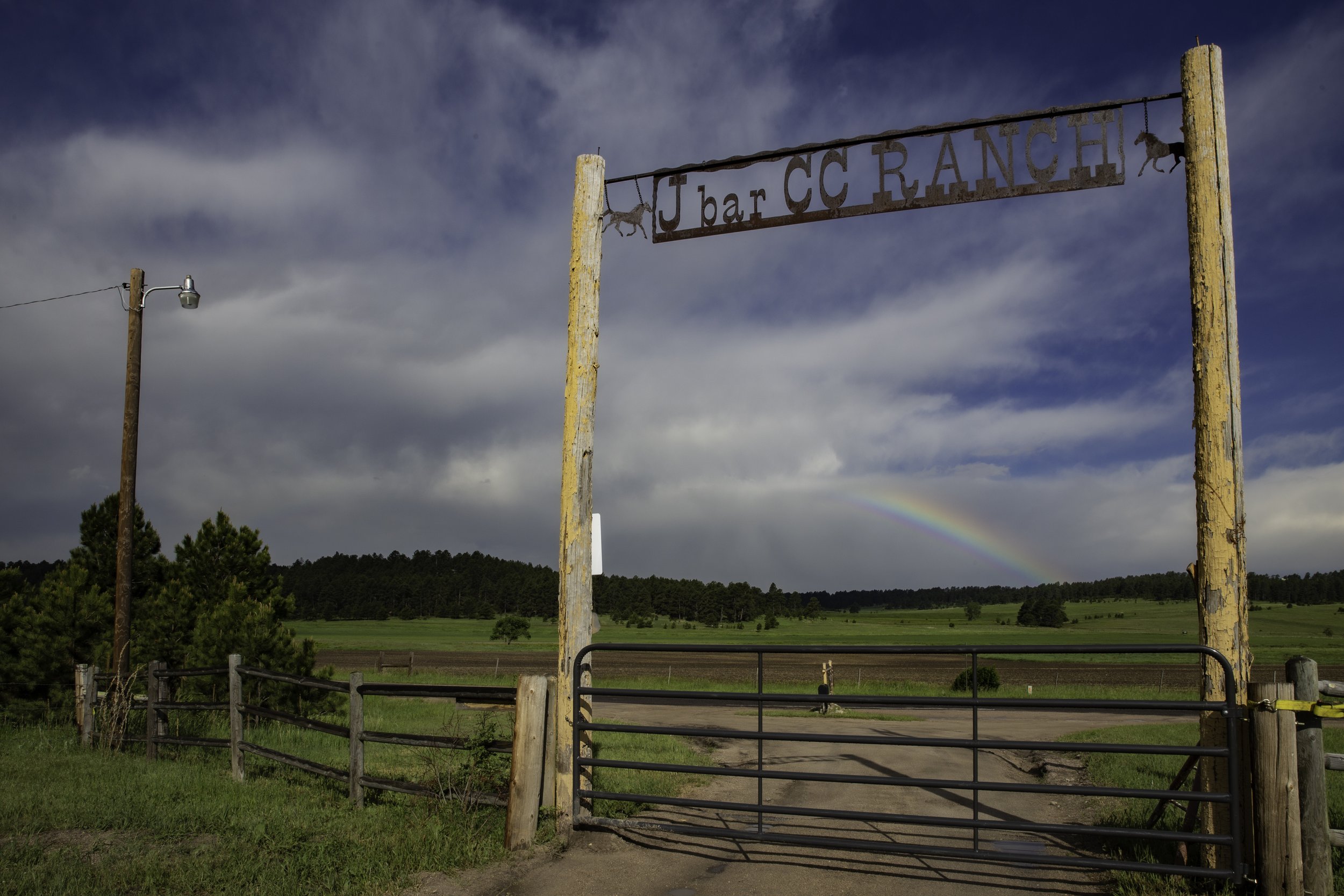 Ranch entrance gate with a wooden sign reading "J bar CC RANCH" across two tall wooden posts, a dirt road leading into open green fields, and a rainbow in the cloudy sky.