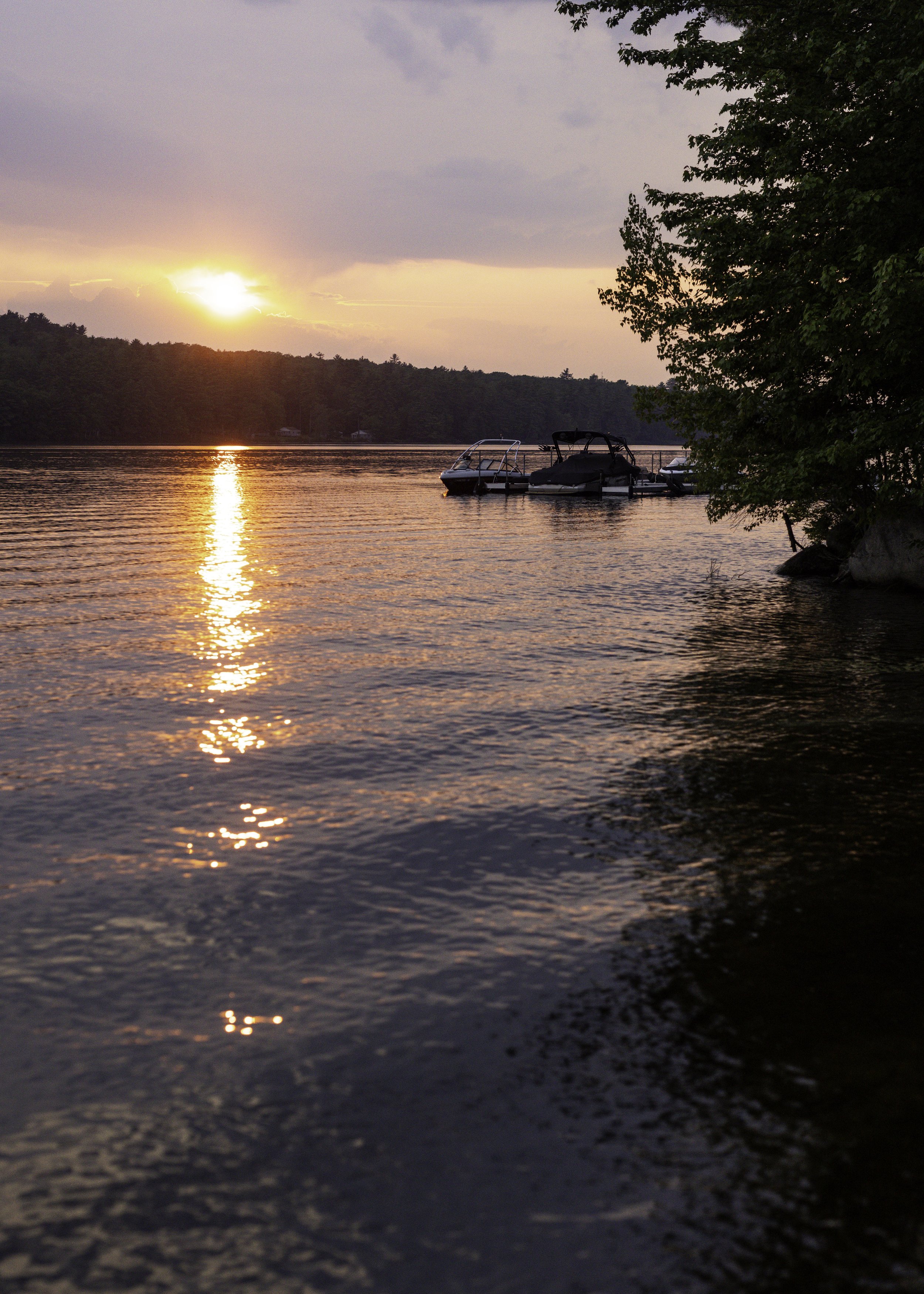 Sunset over a lake with boats docked near the shore, trees on the right side, and the sky with pink and purple clouds.