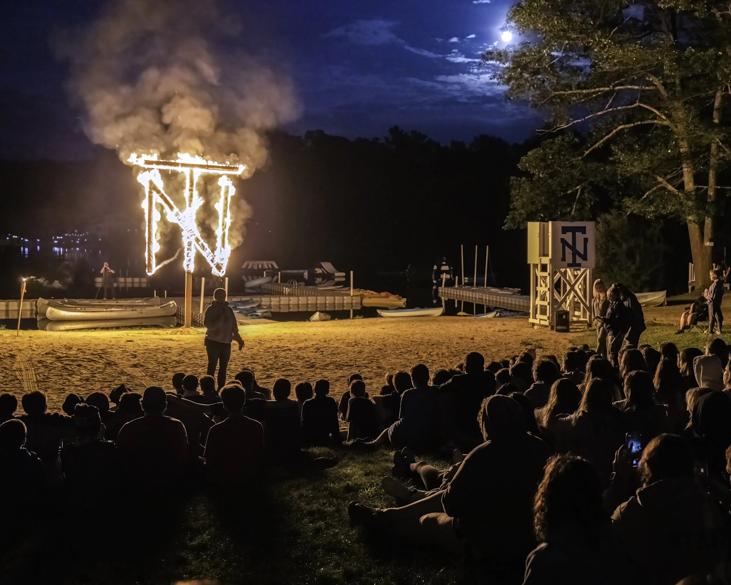 A group of people sitting on grass near a waterfront at night, watching a burning torch sculpture in the shape of a letter 'N'. The scene is illuminated by the fire and the moonlight, with boats docked at a pier in the background and trees on the rig