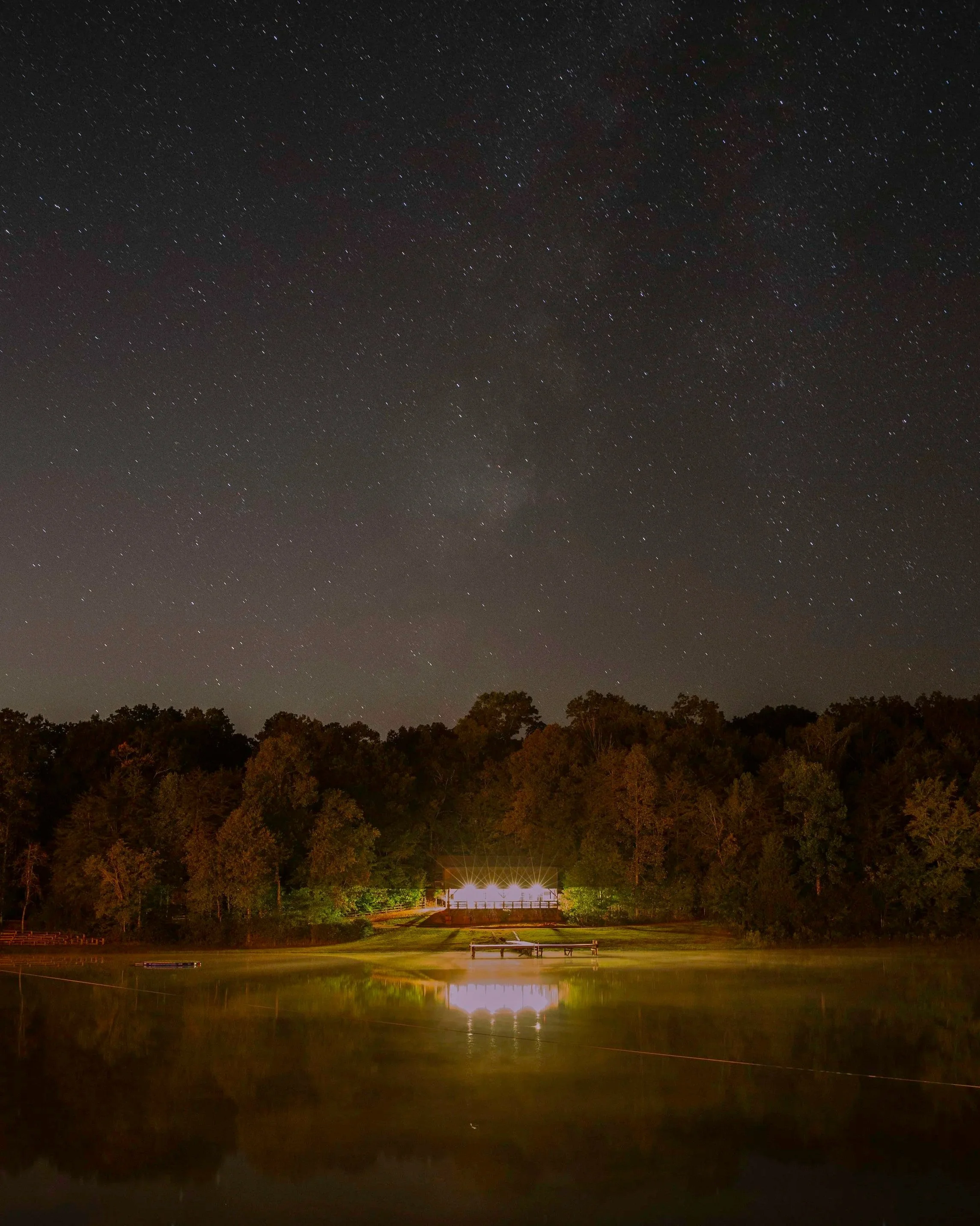 Nighttime scene of a lake with a dock, surrounded by trees, under a starry sky with visible stars and the Milky Way, and a lit building or pavilion near the trees reflecting on the water.