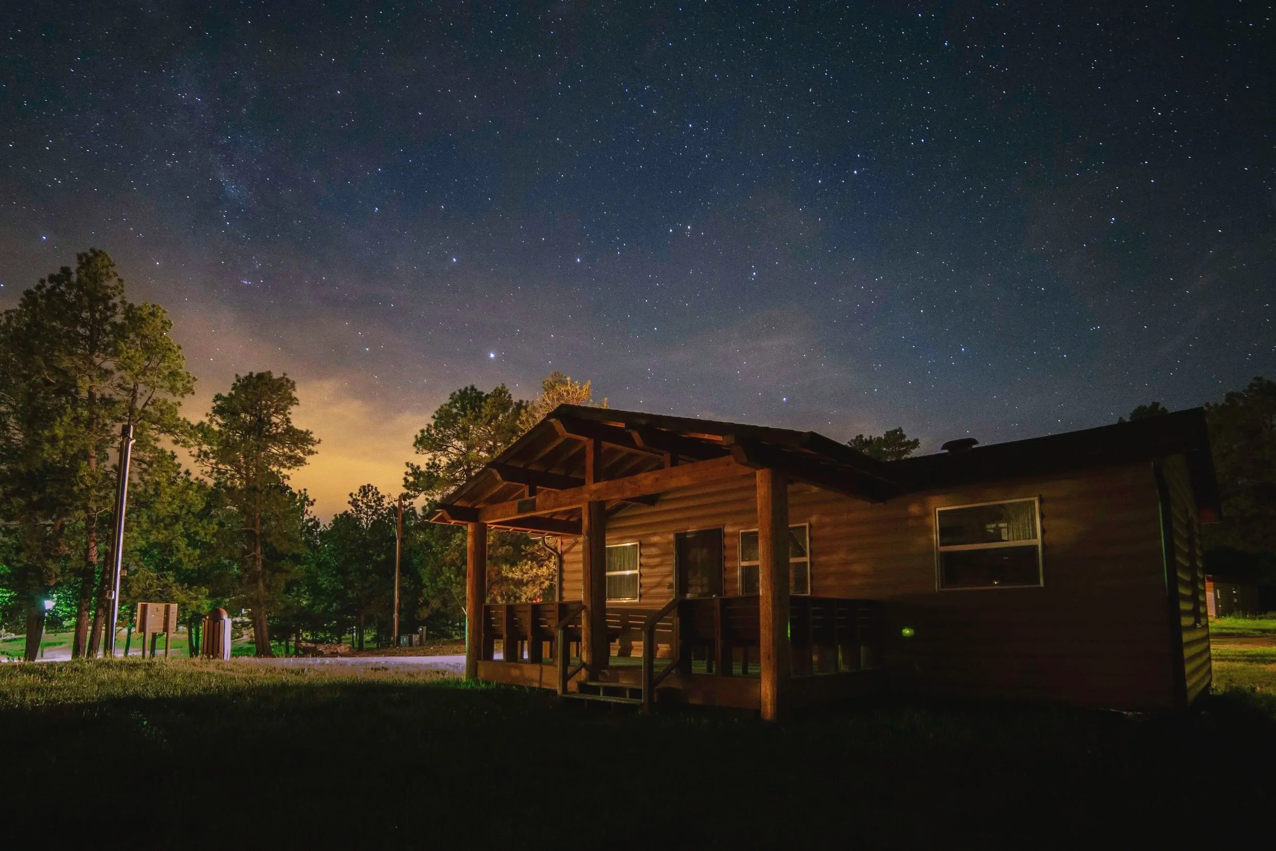 Nighttime scene with a wooden house surrounded by trees, under a starry sky with the Milky Way visible.