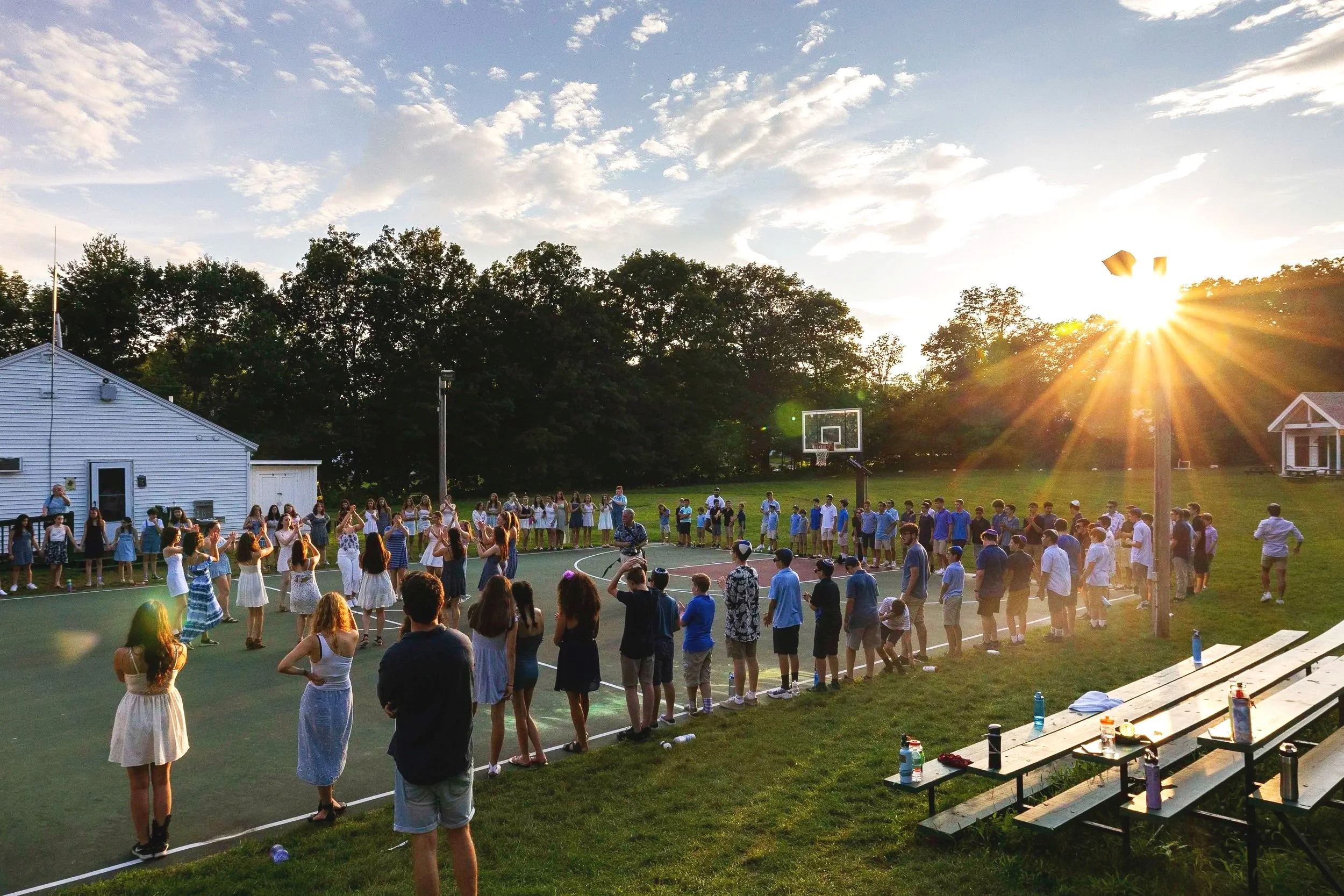 A large group of children and teenagers standing in a circle around the basketball court at sunset, engaging in an outdoor activity or dance. There are benches and tables with water bottles nearby, and a building and trees in the background.