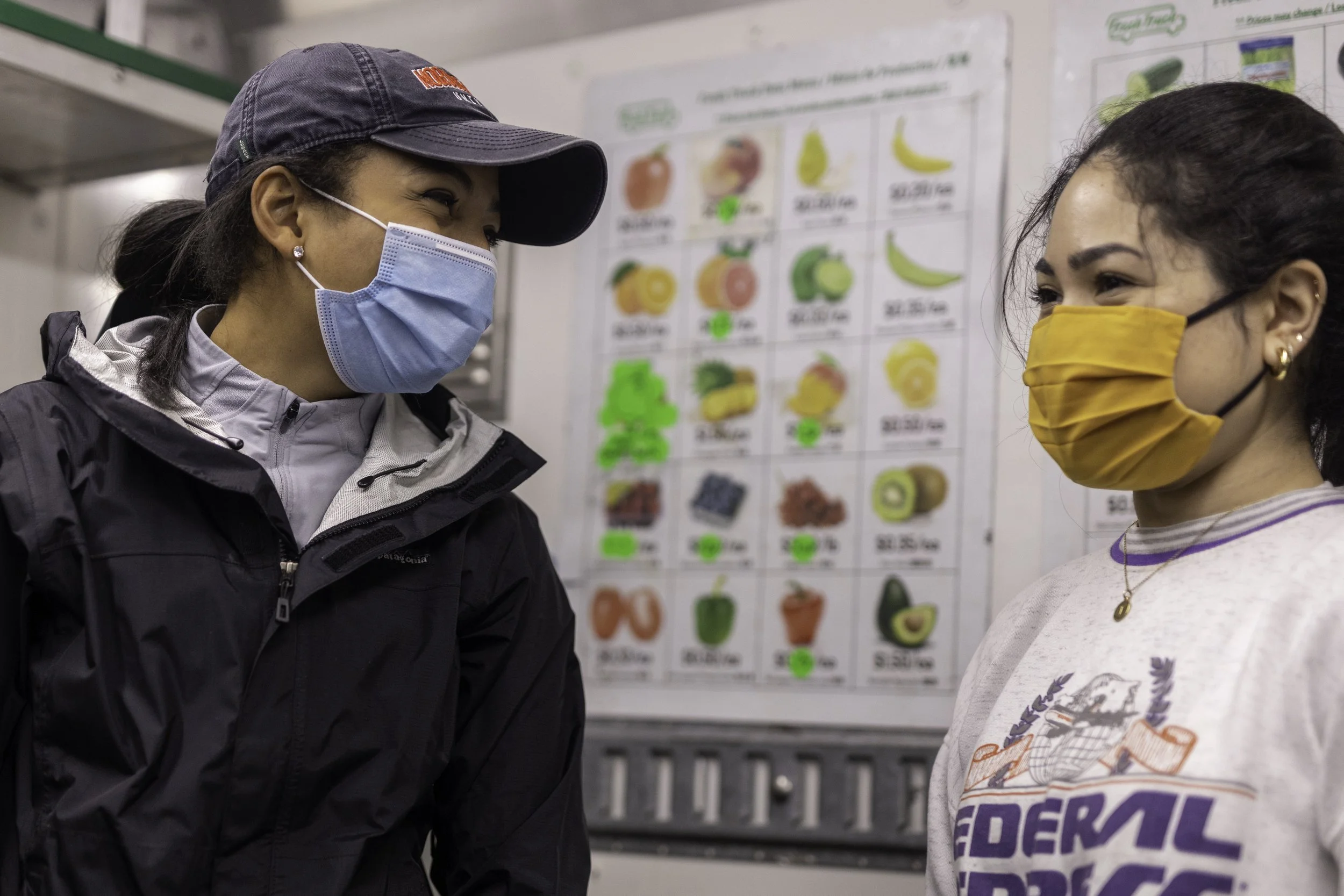 Two women wearing face masks and conversing in front of a fruit menu board at a grocery store or market. The woman on the left has dark hair, a navy cap, and a black jacket. The woman on the right has dark hair pulled back and a mustard-colored mask,