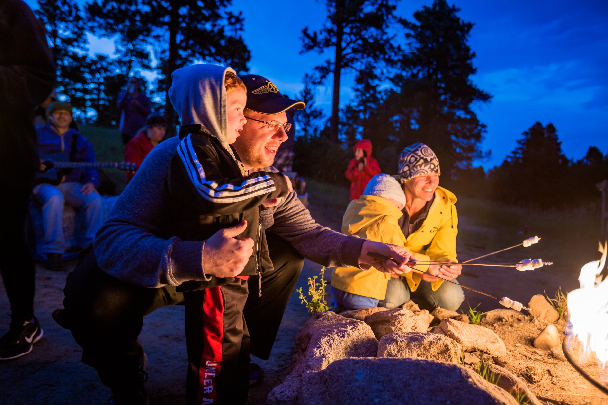 People roasting marshmallows over an outdoor campfire at dusk, with trees and a blue sky in the background.