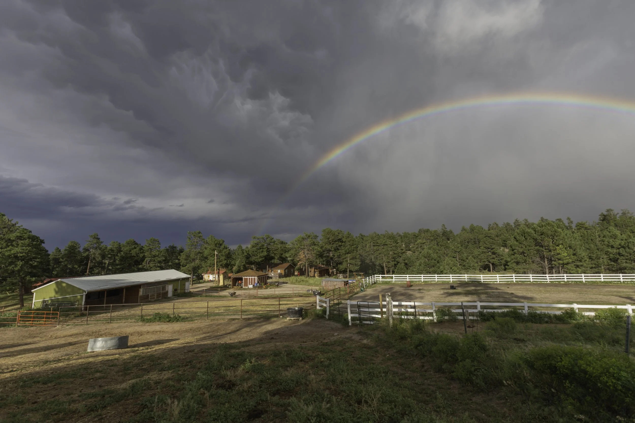 A rural farm under a stormy sky with a rainbow arching over the trees and farm buildings, including a green barn, surrounded by fenced fields.