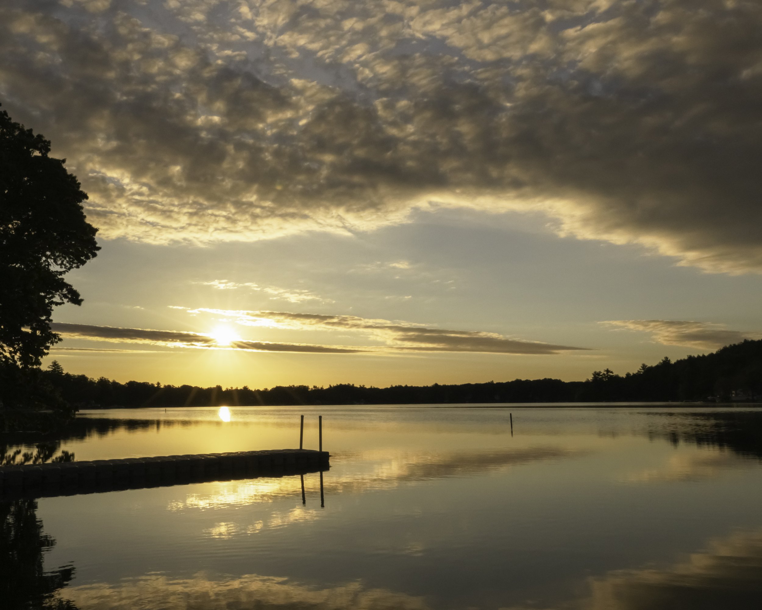 Sunset over a calm lake, with reflective water and a pier on the left side, under a partly cloudy sky.