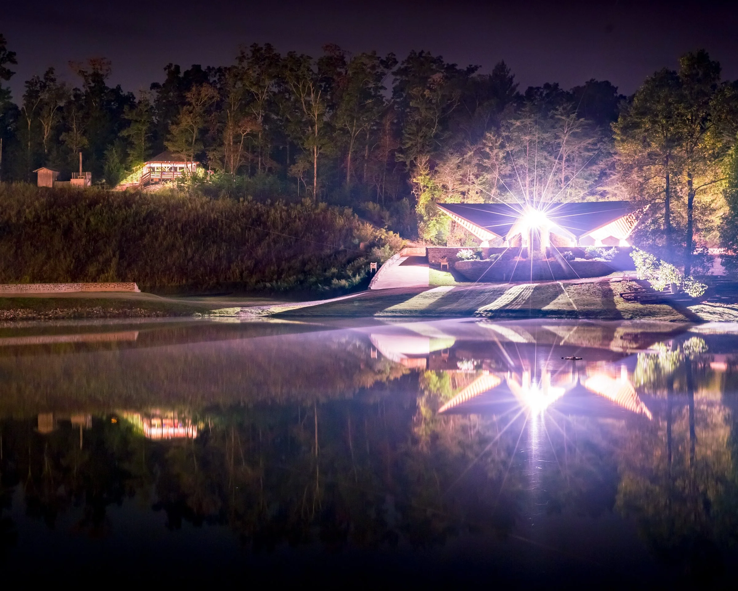 Night scene of a landscaped park with a illuminated pavilion and trees reflected on a calm body of water.