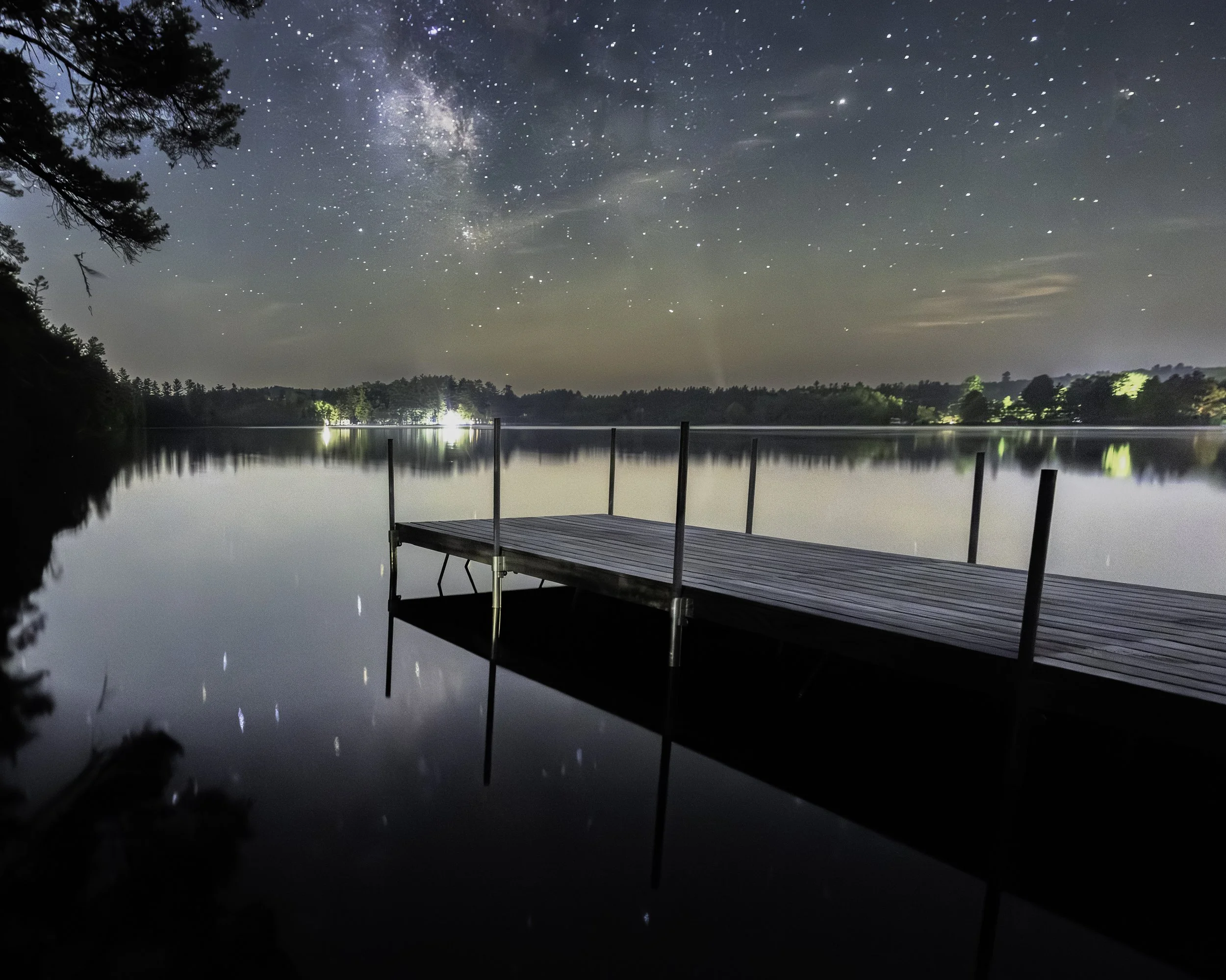 A wooden dock extends into a calm lake at night, with a star-filled sky and the Milky Way visible, and faint lights from distant houses reflecting on the water.