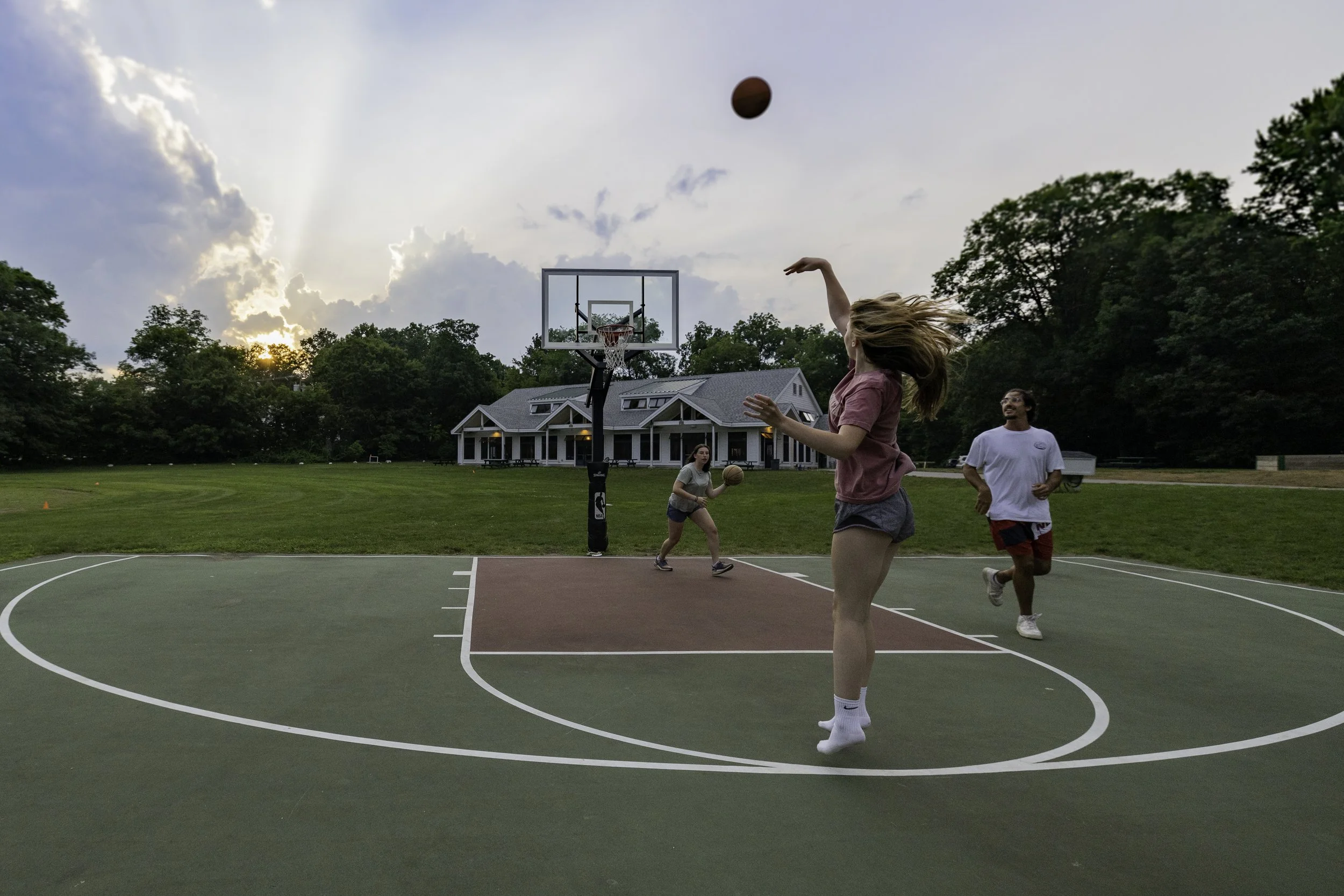 People playing basketball on an outdoor court during the evening, with trees and a house in the background.