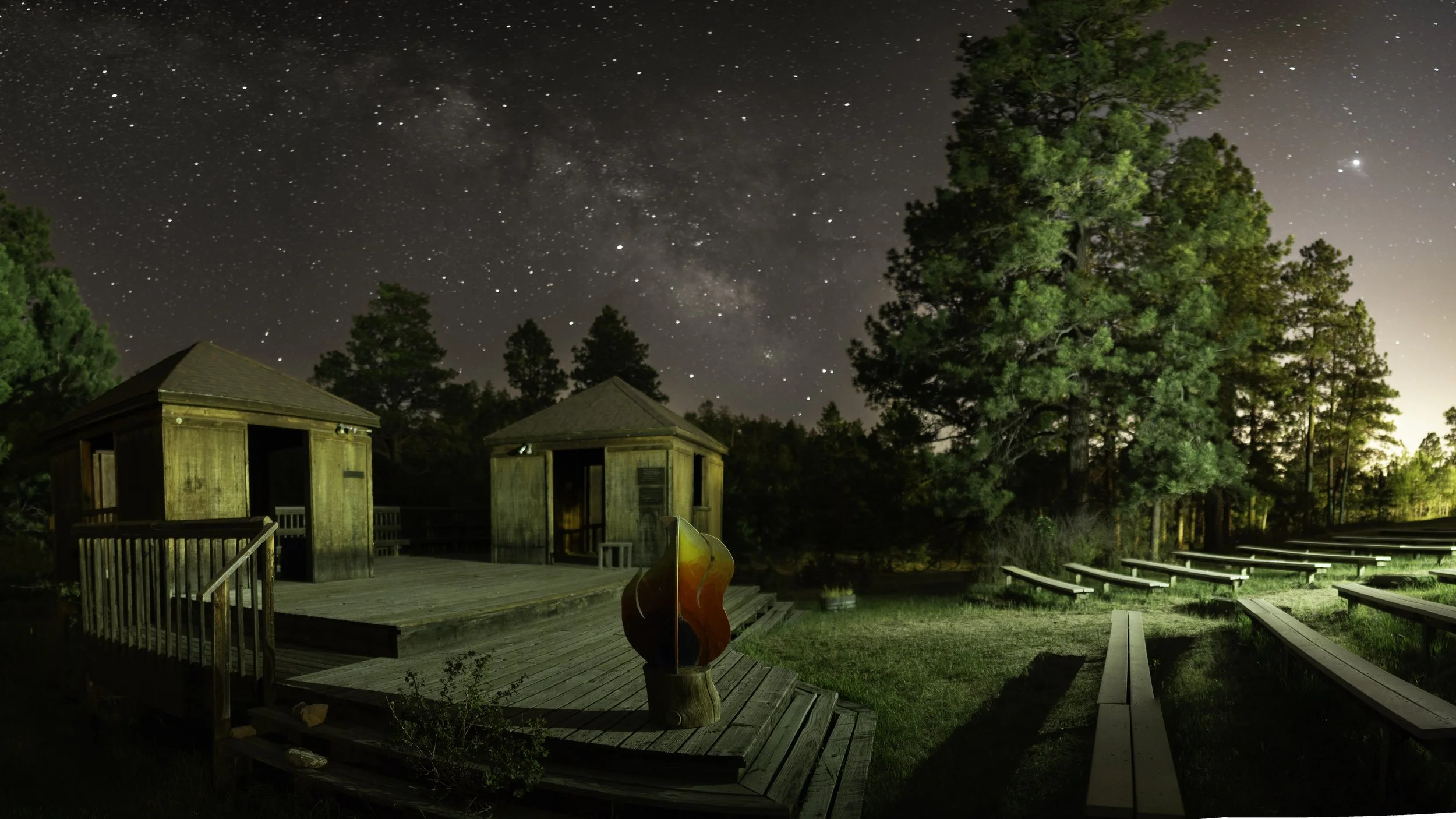 Nighttime scene of a wooden outdoor amphitheater with steps and seating, two small wooden buildings, and tall trees, under a starry sky with the Milky Way visible.