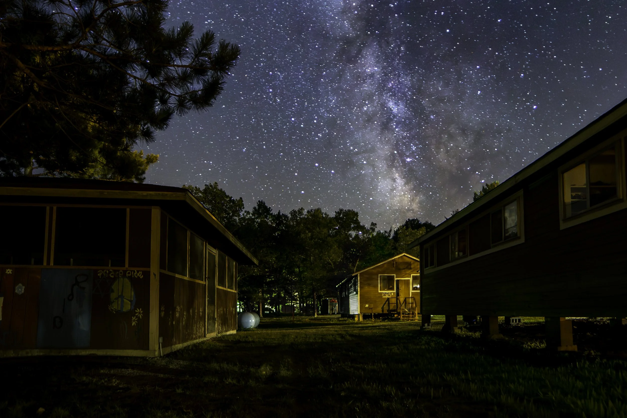Nighttime scene of a starry sky with the Milky Way galaxy visible above small cabins and trees.