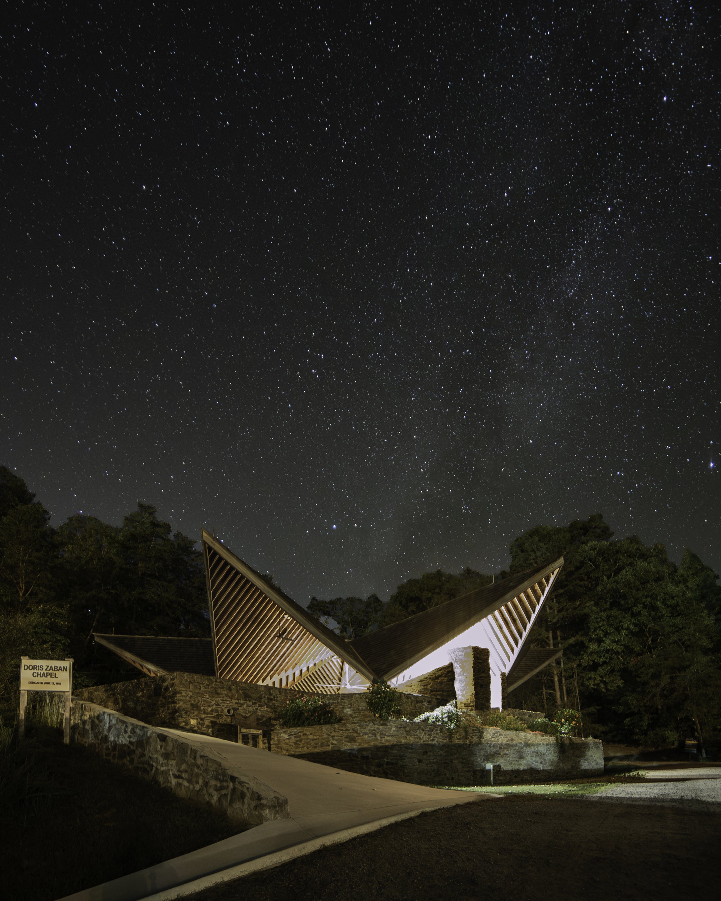 Night sky filled with stars above a modern chapel with angular roof design, surrounded by trees and a landscaped area.