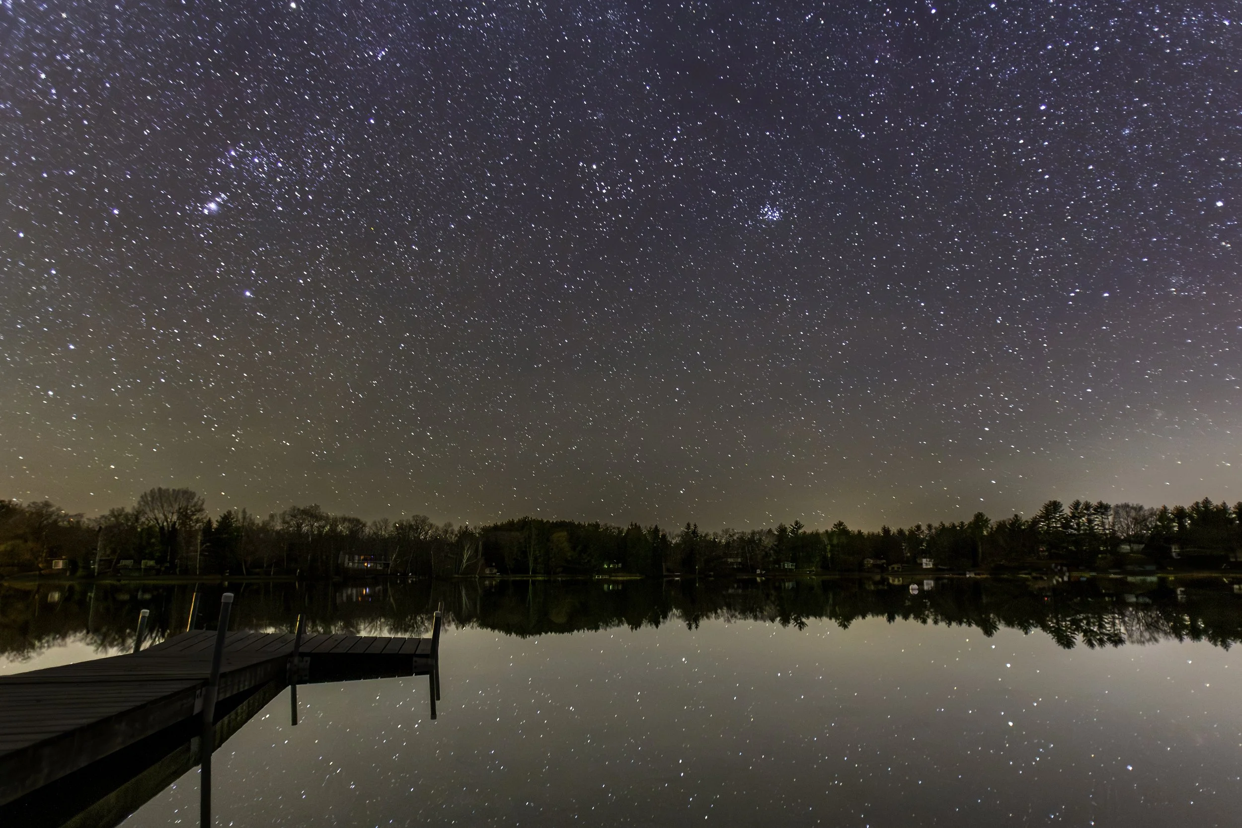 Night sky filled with stars reflected on a calm lake, with a wooden dock extending into the water in the foreground, and a tree-lined shoreline in the distance.