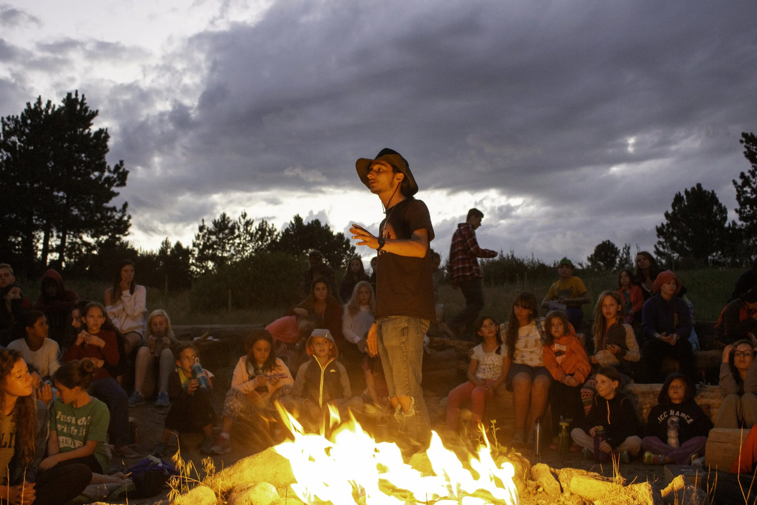 A man wearing a large hat and dark clothing stands in front of a campfire during dusk, surrounded by a group of children and teenagers sitting and watching outdoors in a natural area with trees and cloudy sky.