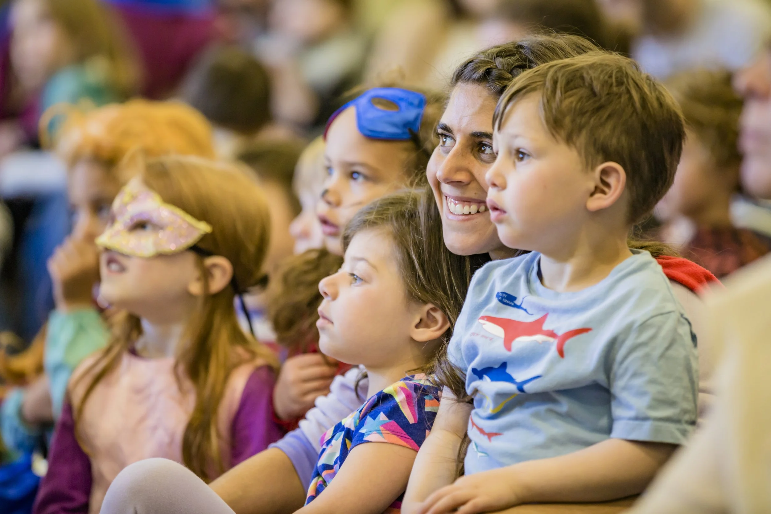A group of children and a woman sitting in an audience, attentively watching a performance. One girl wears a pink mask, and a boy wears a blue whale shirt. The woman, with long hair, smiles while the children watch.