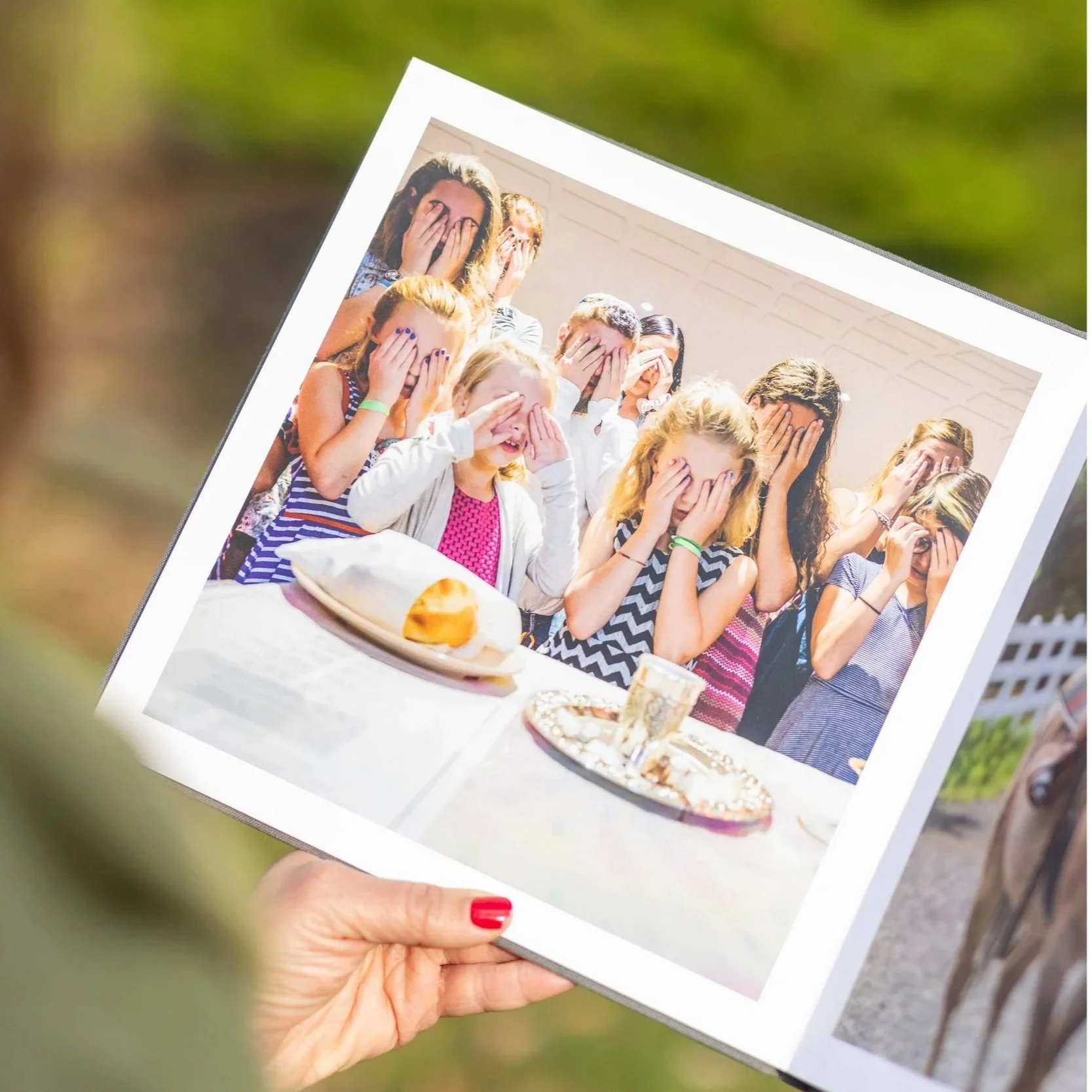 A hand holding a printed photo of a group of young people at a party, all covering their faces with their hands, with snacks on the table in the foreground.