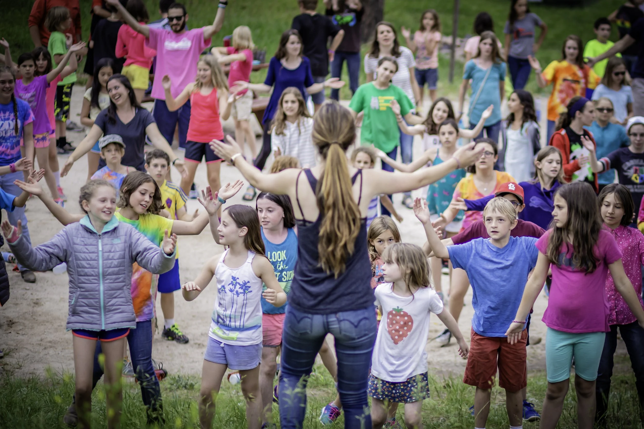 A woman leading a group of children in an outdoor dance or movement activity in a park or natural setting.