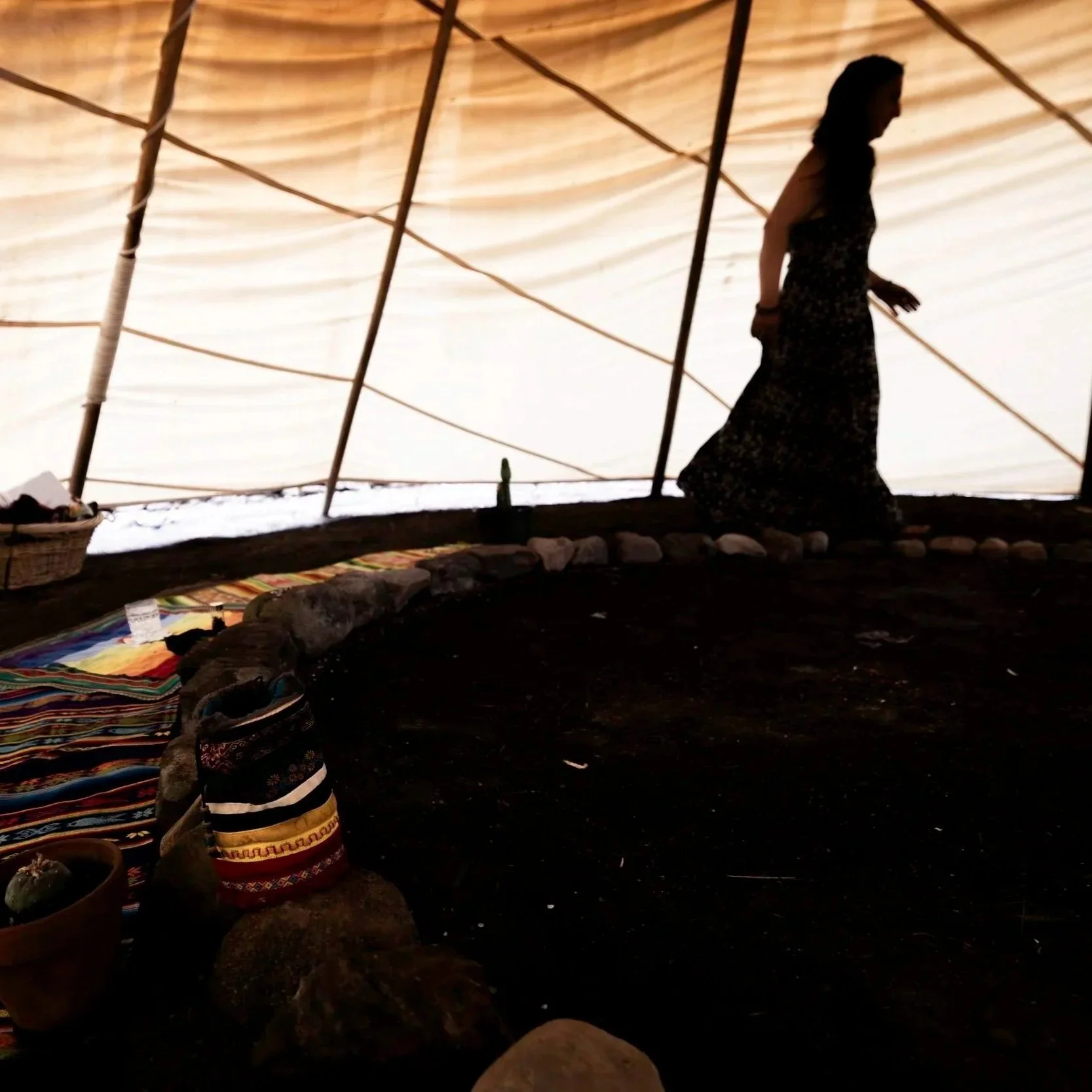 A person walking inside a tent with a wooden framework and a fabric ceiling, sitting on rocky ground with colorful woven rugs.