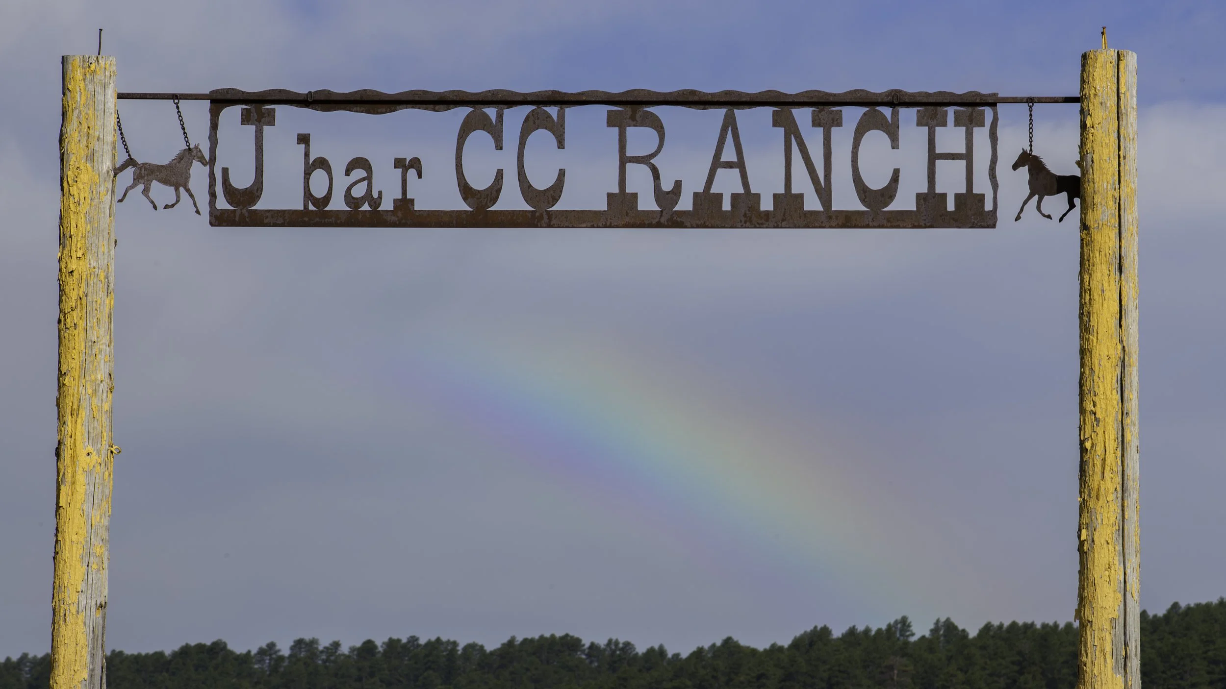 Rusty metal ranch entrance sign reading 'J bar C C Ranch' with two horse-shaped decorations hanging on chains, mounted between two weathered wooden posts with yellow peeling paint, against a background of a cloudy sky and a faint rainbow over a fores