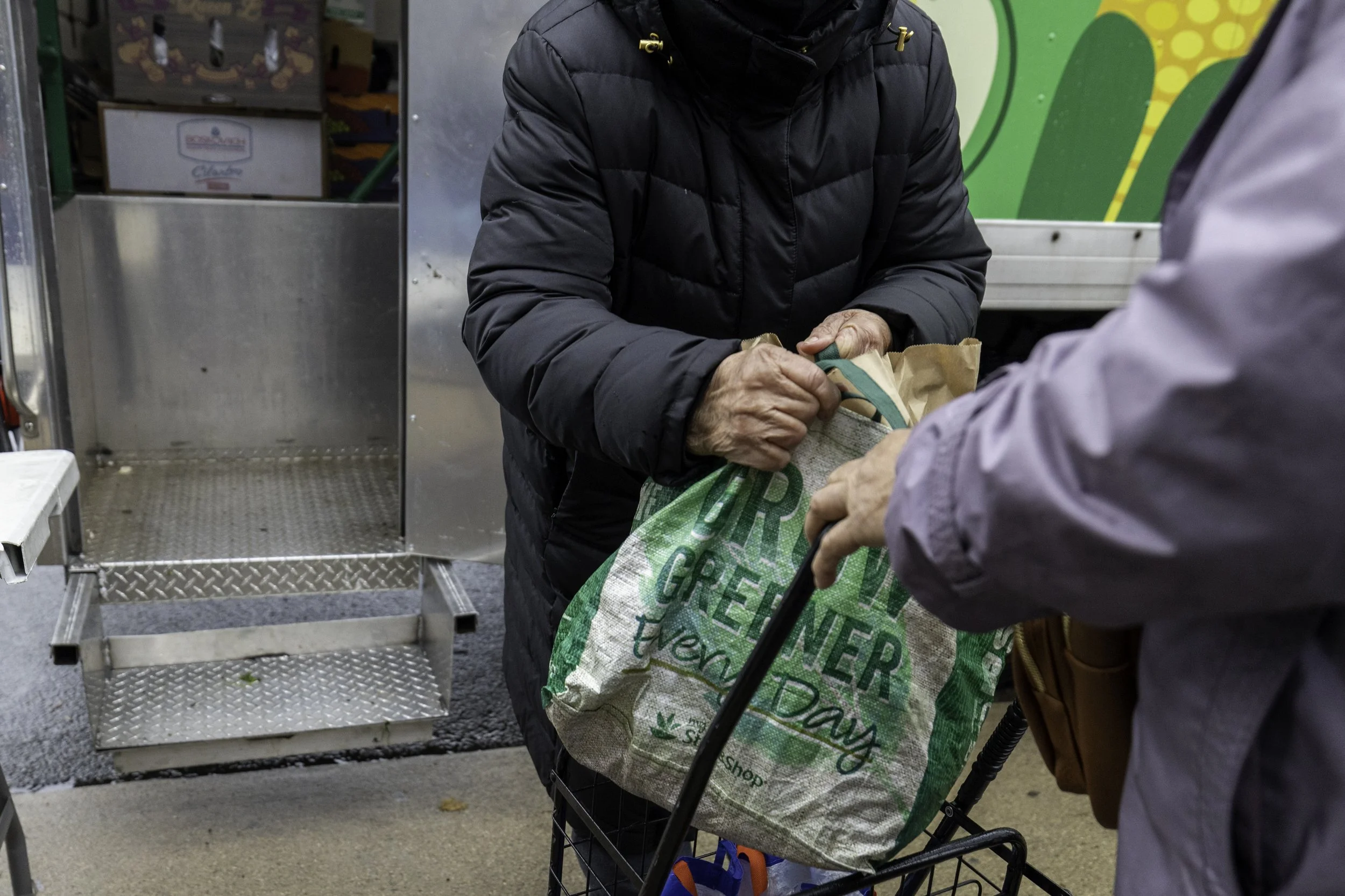Two people exchanging a reusable grocery bag outside a store. One person is wearing a black puffer jacket, and the other is wearing a gray jacket.