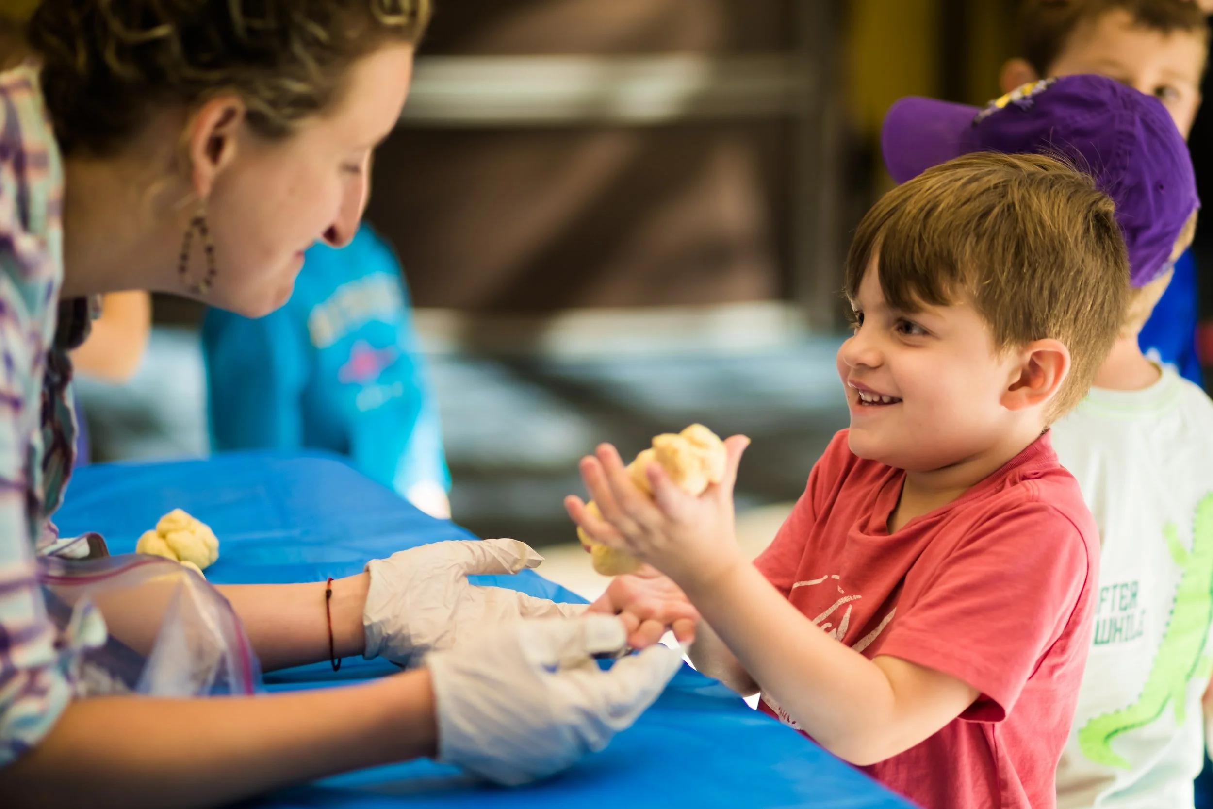 Child receiving a piece of dough from a woman, likely during a baking or cooking activity, with other children in the background.