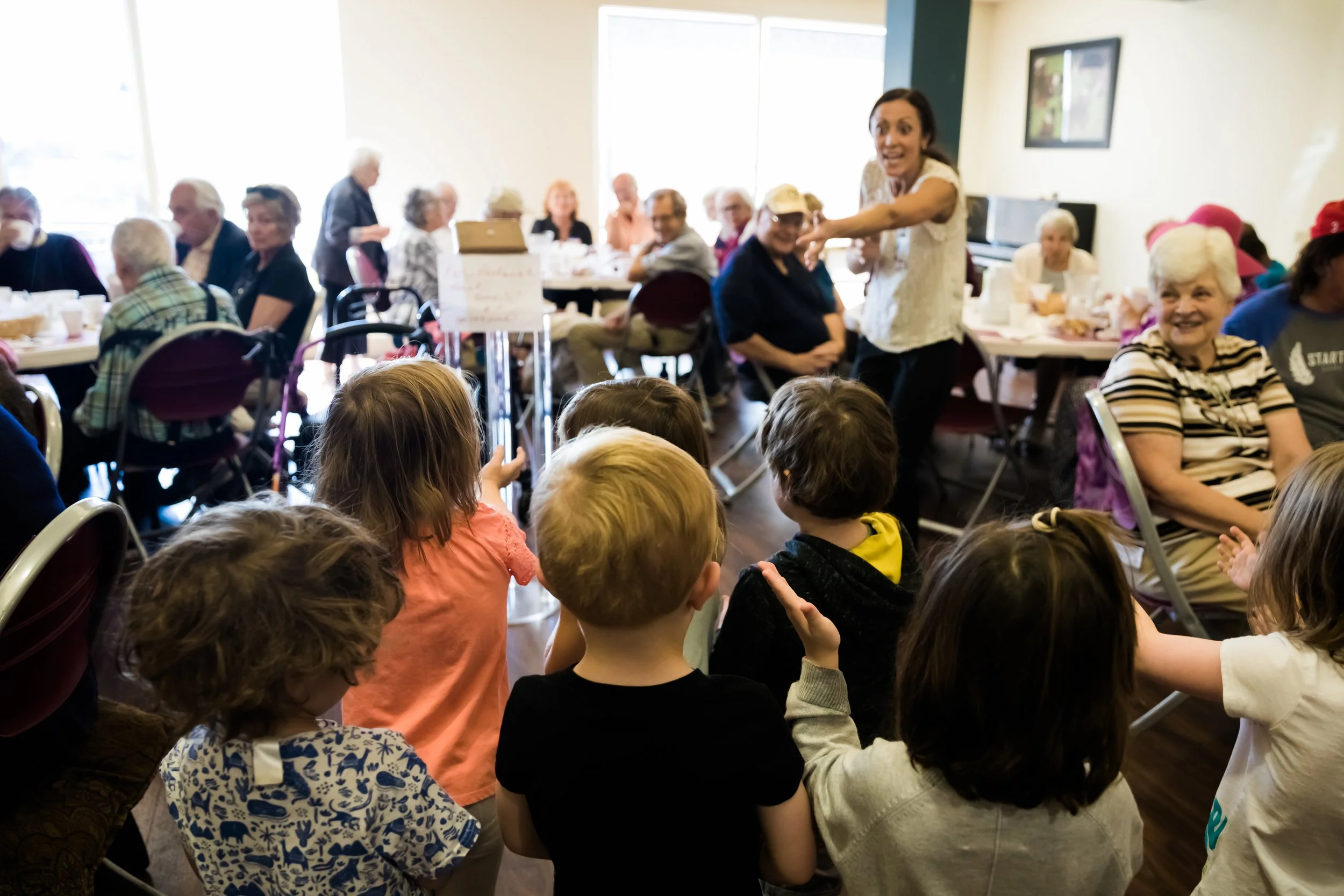 Children and senior citizens gathered in a community room, engaging in a group activity. A woman in a white top is talking to the children, who are sitting and raising their hands. Seniors sit at tables in the background, some smiling and watching th