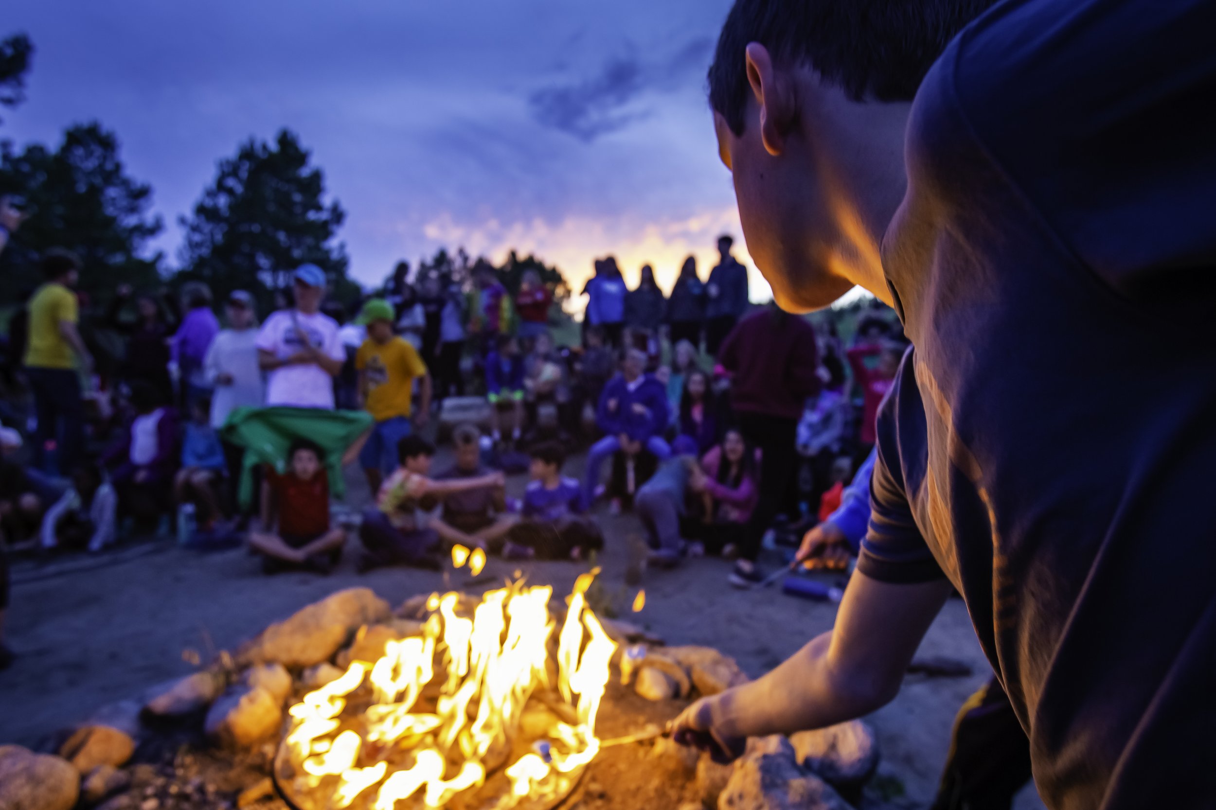 Child tending a campfire surrounded by a crowd outdoors during sunset.
