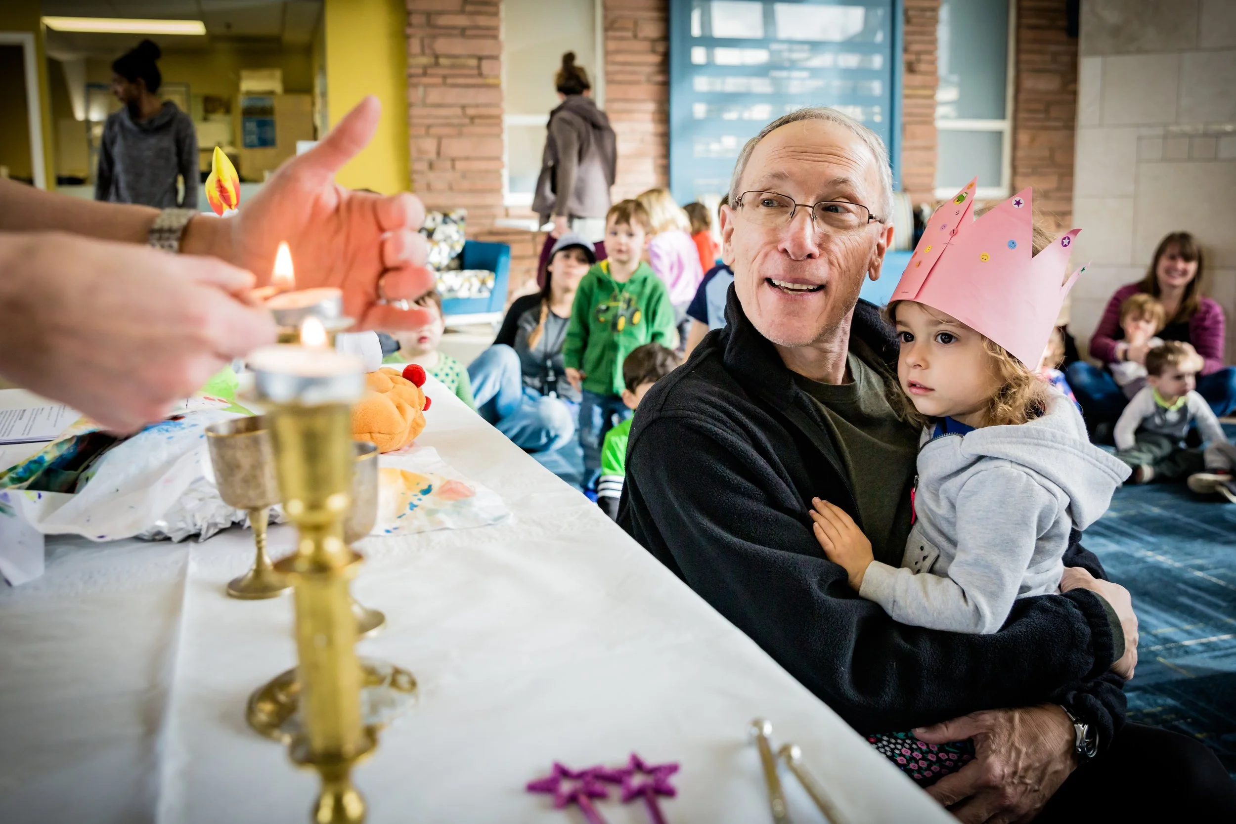 An elderly man is holding a young girl who is wearing a pink paper crown at a birthday celebration with children in the background. A person in the foreground is lighting candles on a birthday cake.