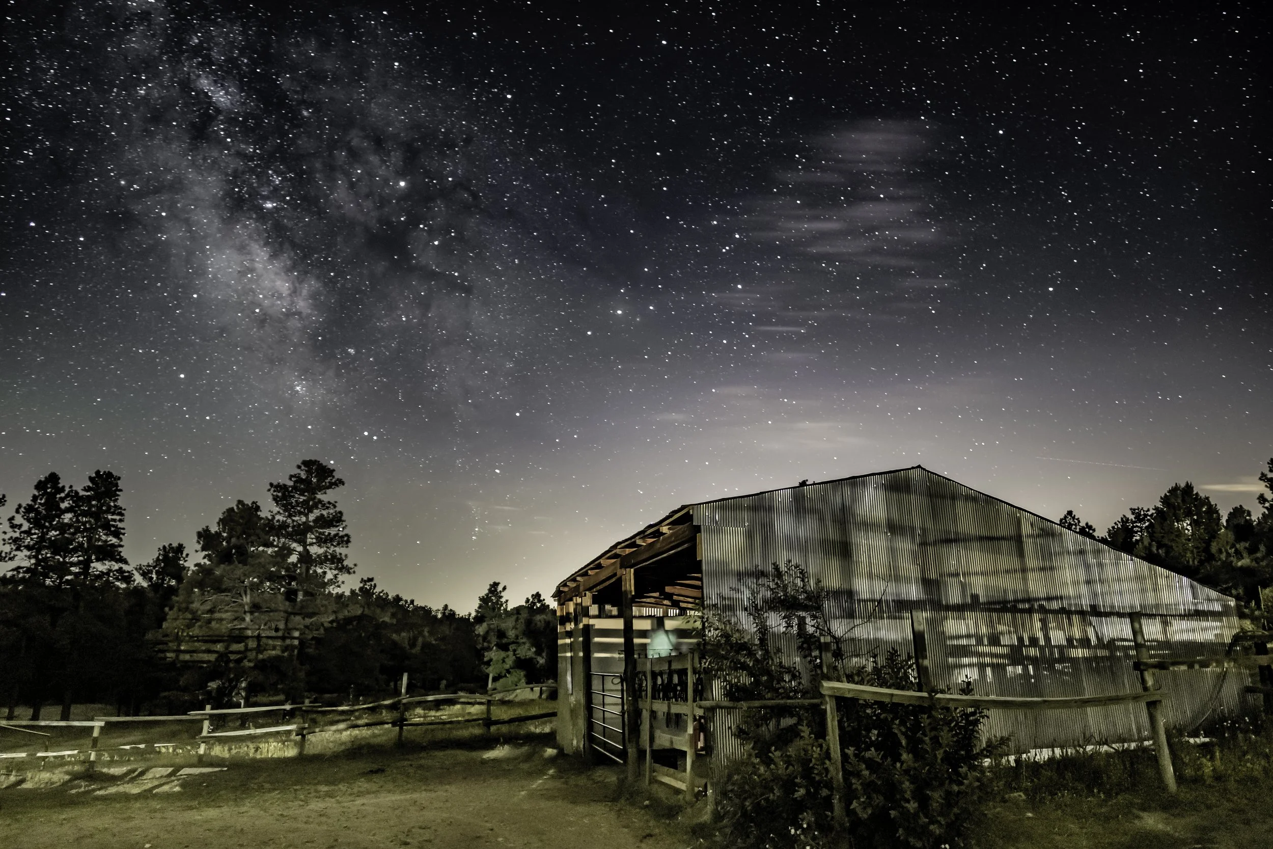 A barn with metal siding illuminated by interior light, set in a rural landscape with trees, under a starry night sky featuring the Milky Way galaxy.