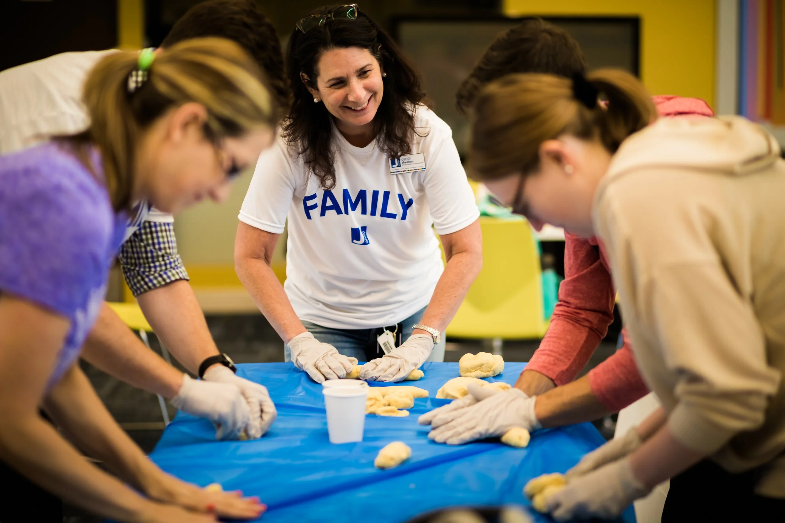 A group of people, including a smiling woman wearing a white t-shirt with the word 'FAMILY' on it, gathered around a table working with dough. All are wearing gloves, and the scene is indoors in a brightly lit room.