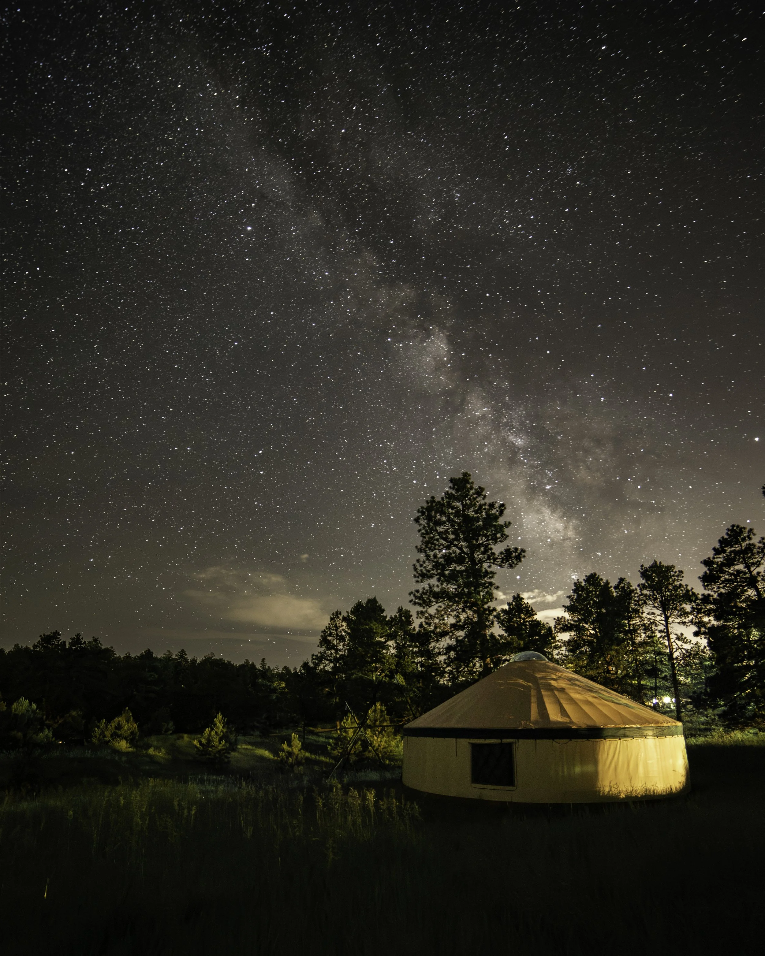 A night sky filled with stars and the Milky Way galaxy, with a small illuminated yurt in a forested area.