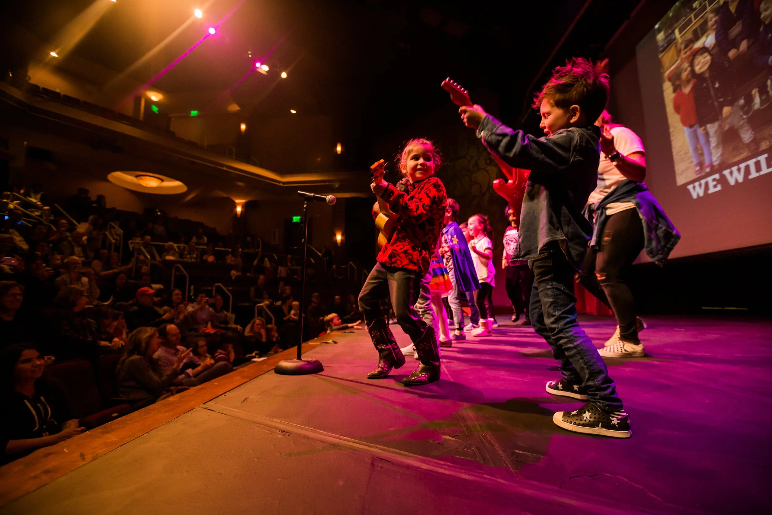 Children performing on stage during a concert or talent show, with an audience watching from their seats.