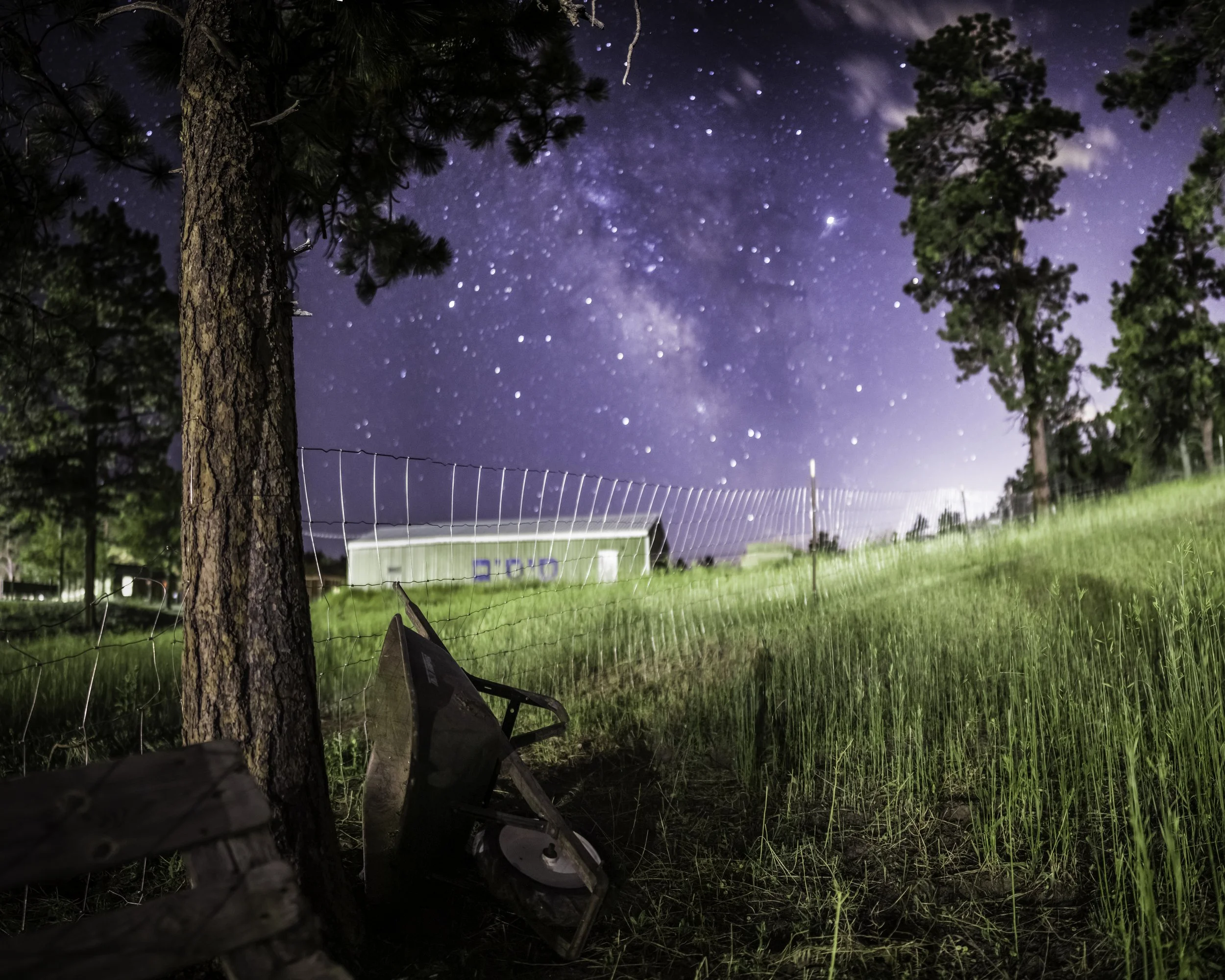 Nighttime scene with starry sky and the Milky Way galaxy, tall trees, a grassy field, and a wooden fence. A wheelbarrow leans against a tree in the foreground.
