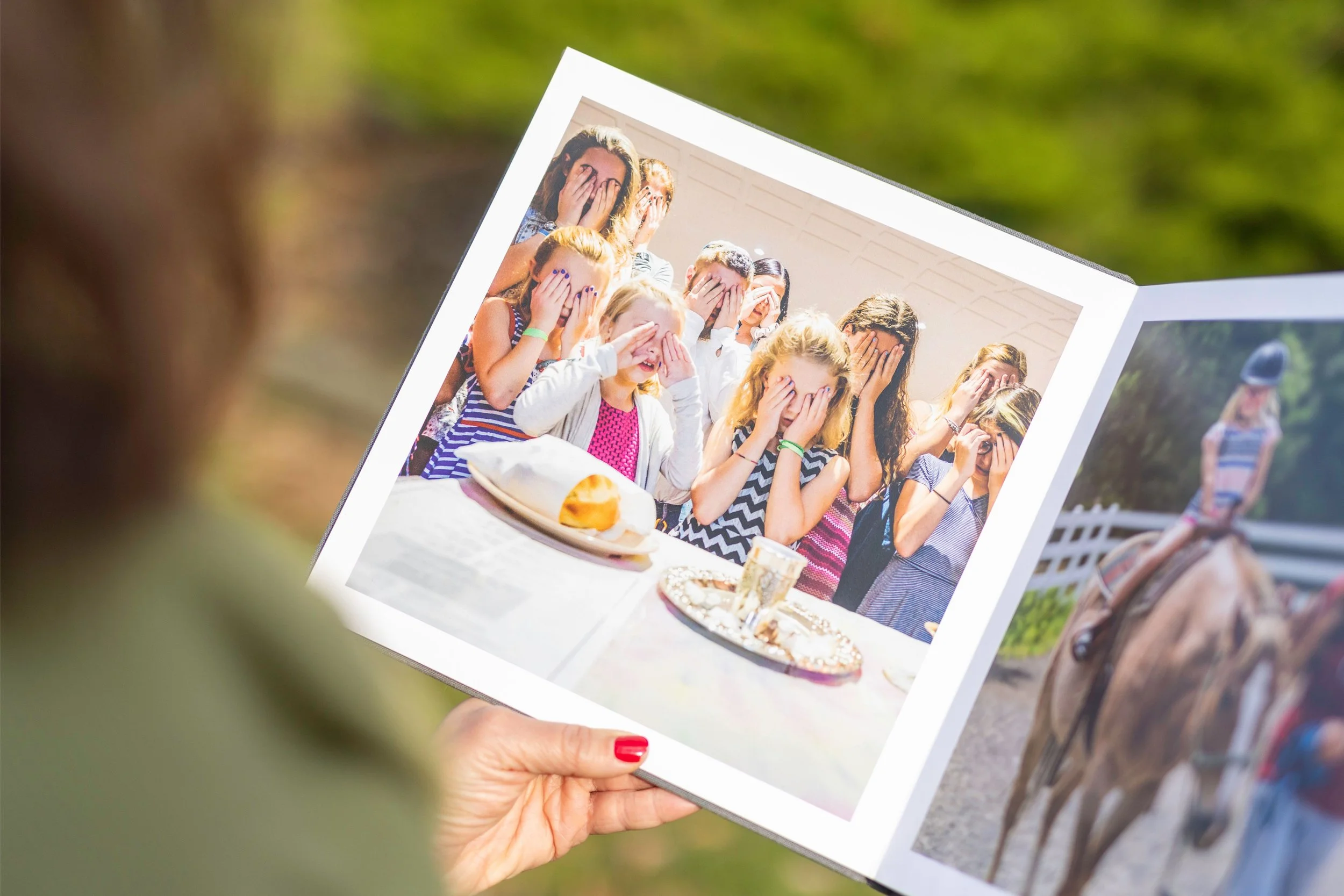 A person holding a photo of a group of people covering their faces with their hands, sitting outdoors at a table with food, surrounded by a blurred green background.