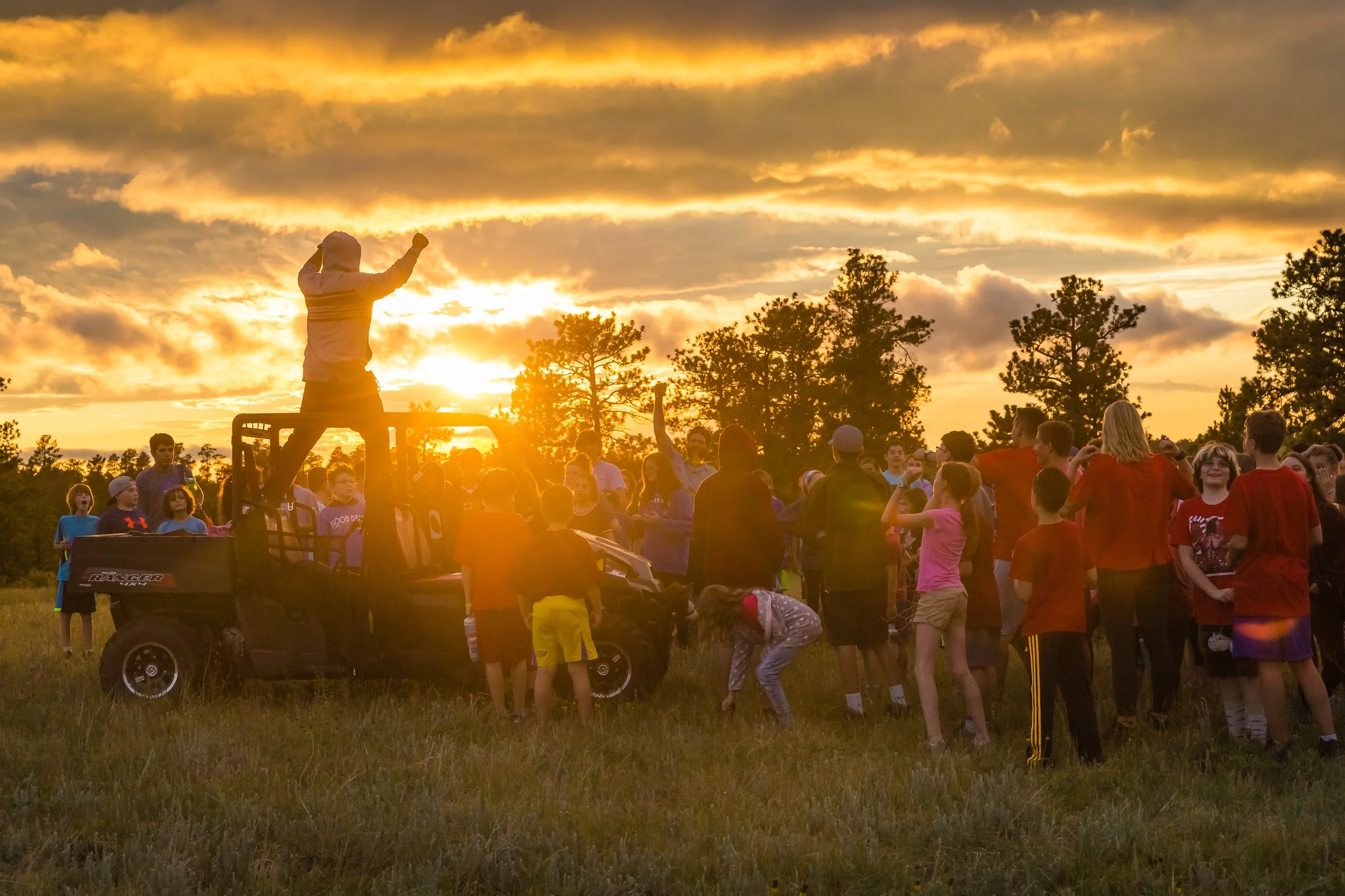Children and adults gathering outdoors at sunset with a person standing on a vehicle ledge, raising one arm, while others are dancing or watching.