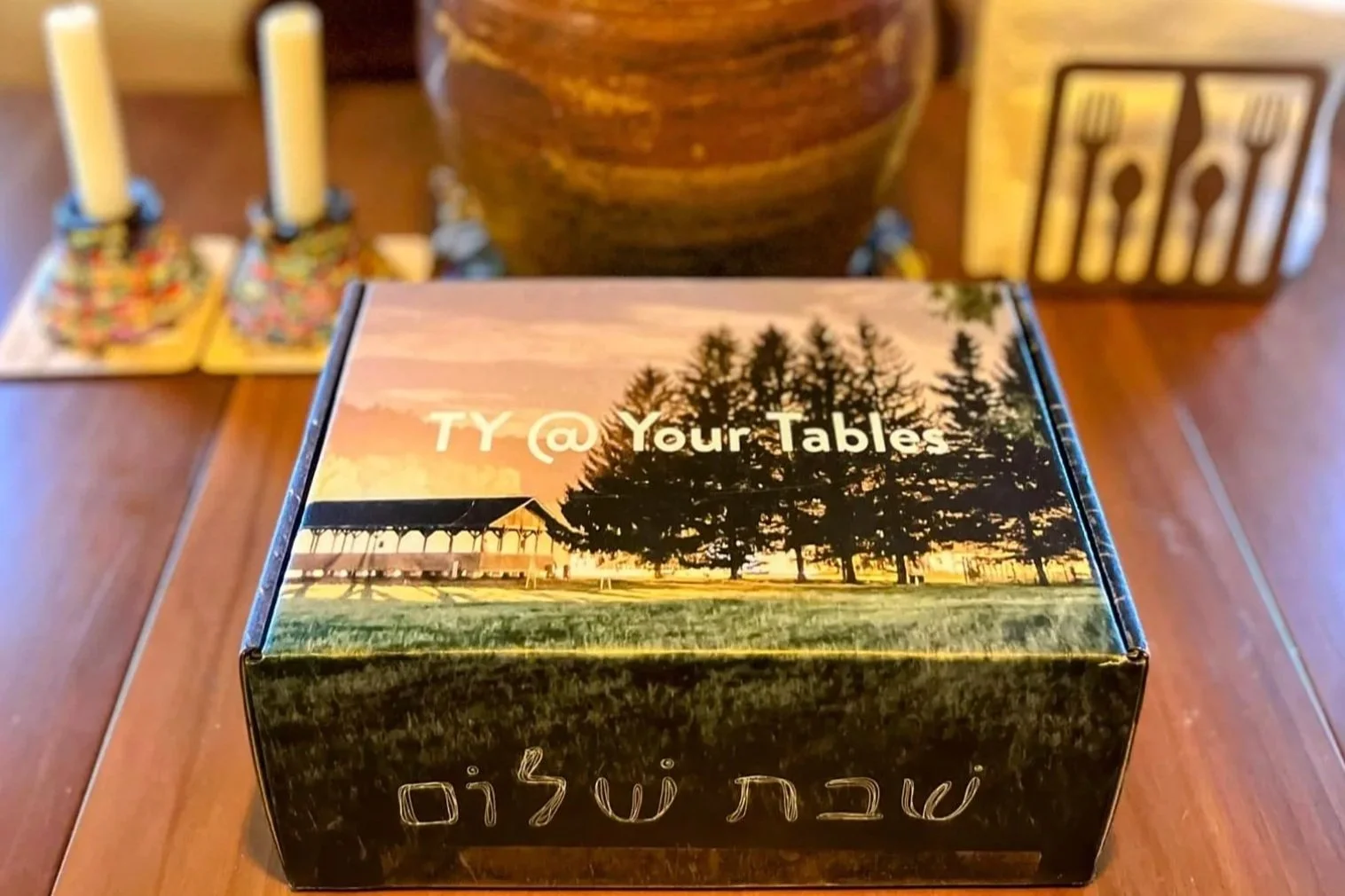A box with a sunset scene and trees on the lid, labeled 'TY @ Your Tables'; background shows candles, a large jar, and utensils.