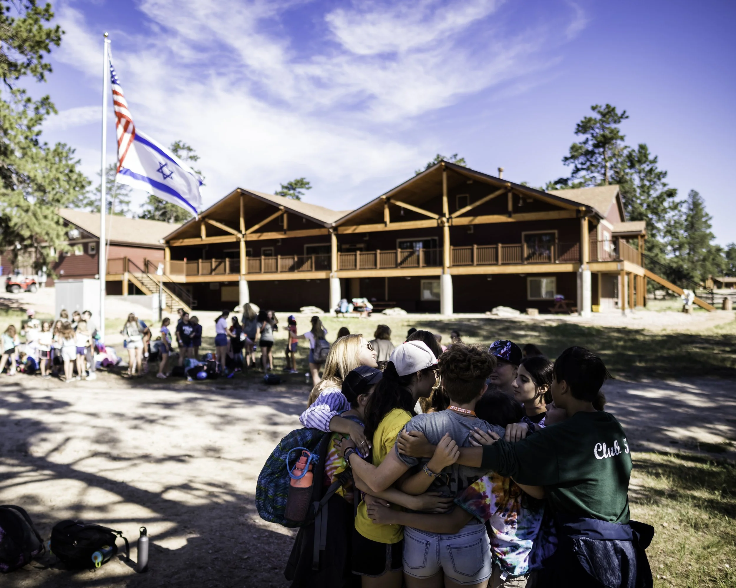 Group of children and campers hugging in front of a wooden lodge at a summer camp, with flags and trees surrounding the area on a sunny day.